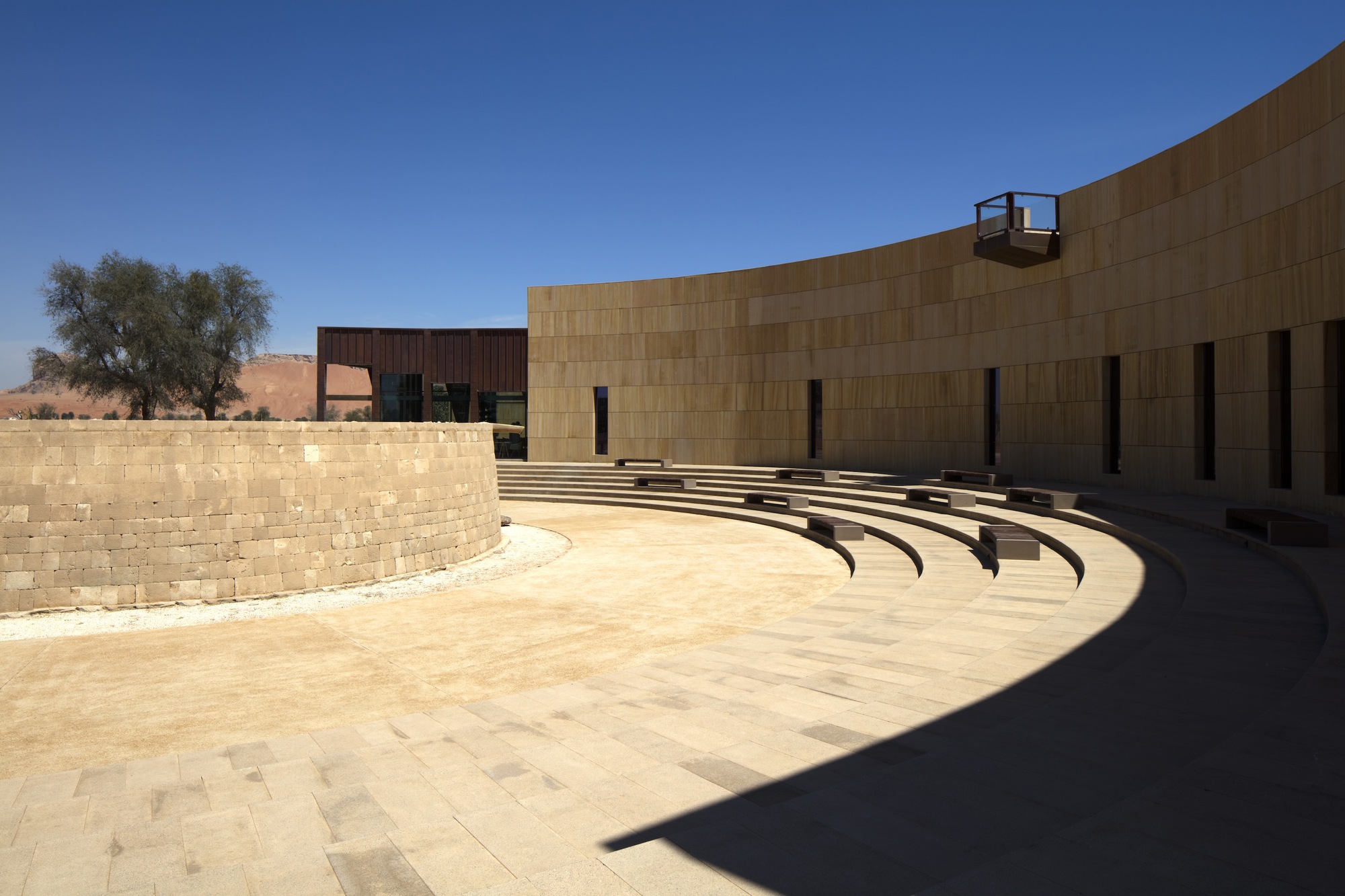 Outdoor amphitheater with curved stone seating tiers and rammed earth walls under a clear blue sky
