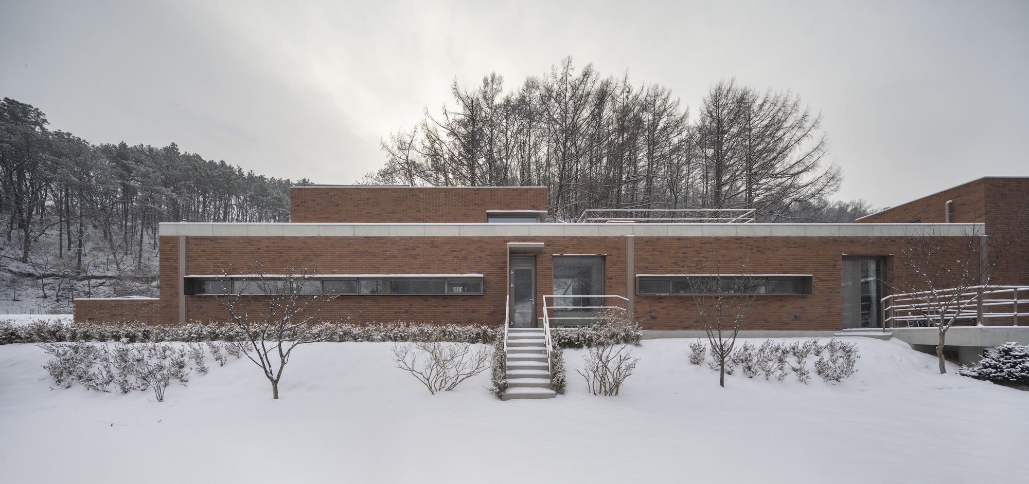 Horizontal brick facade with ribbon windows and front stair in a snow-covered garden beneath winter trees