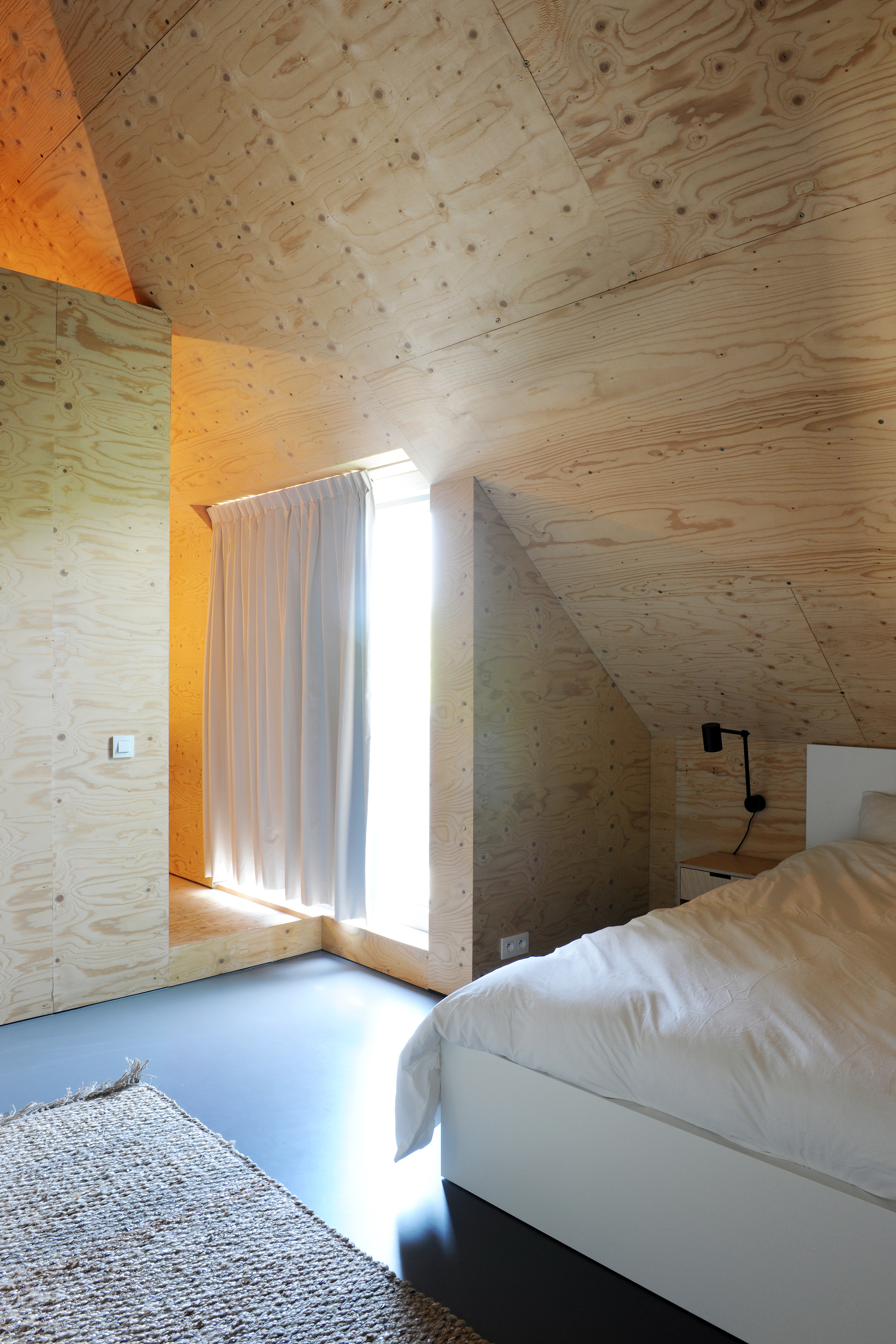 Bedroom with angled plywood ceiling and walls, bright sunlight streaming through curtained opening beside the bed