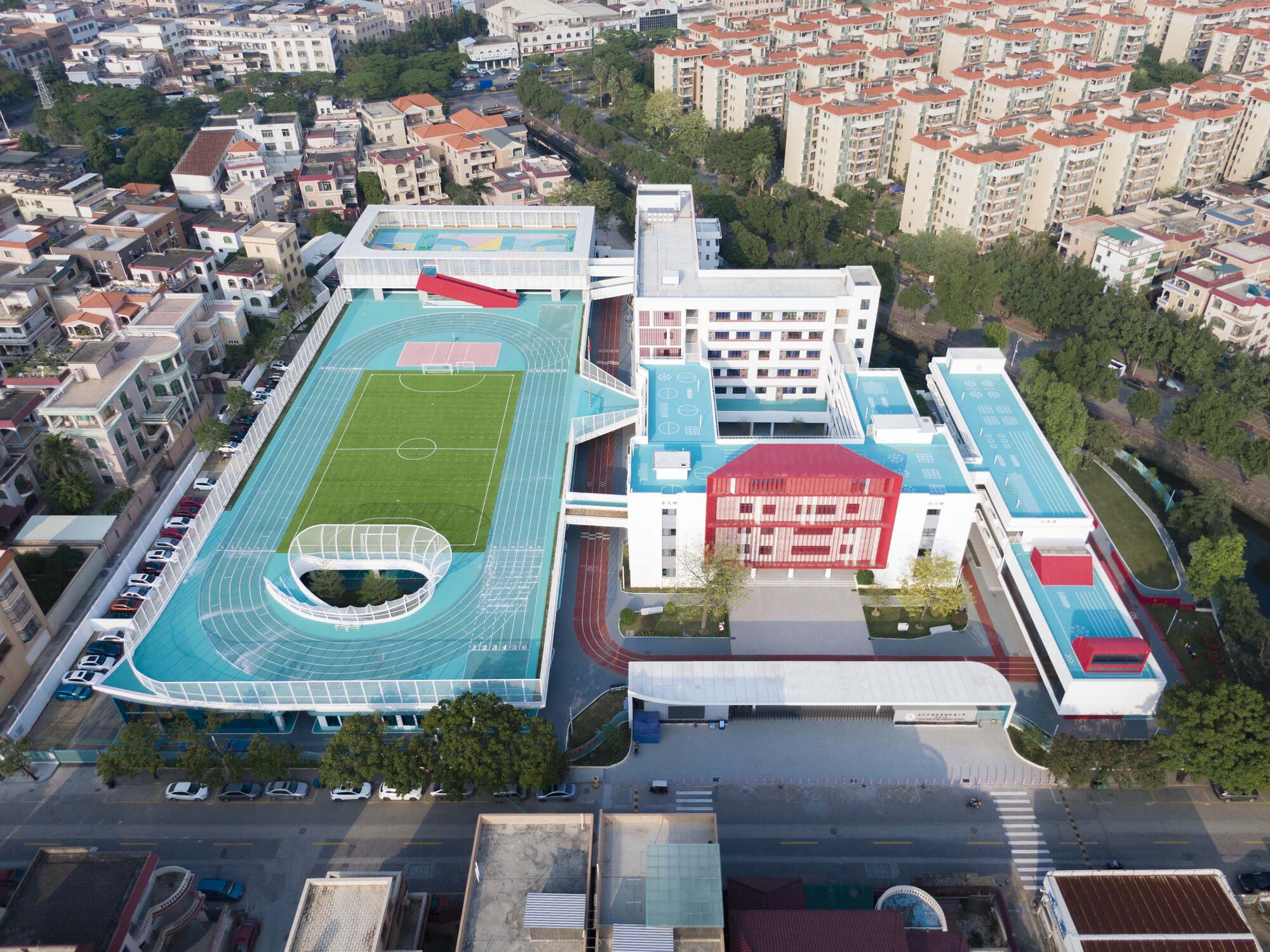 Aerial view of the school campus with turquoise running track, soccer pitch and white buildings set in dense residential neighborhood