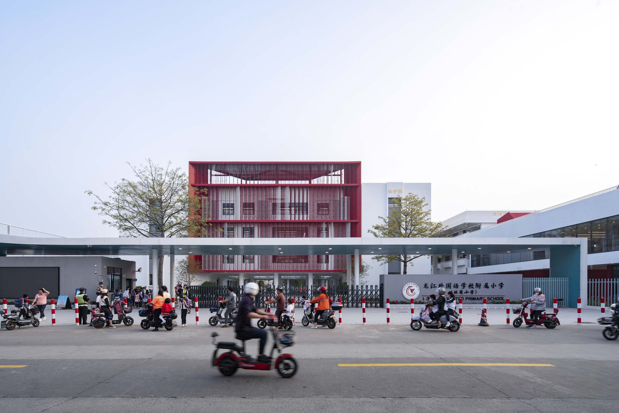 Street view of the red louvered facade and elevated pedestrian bridge with scooters passing by at dusk