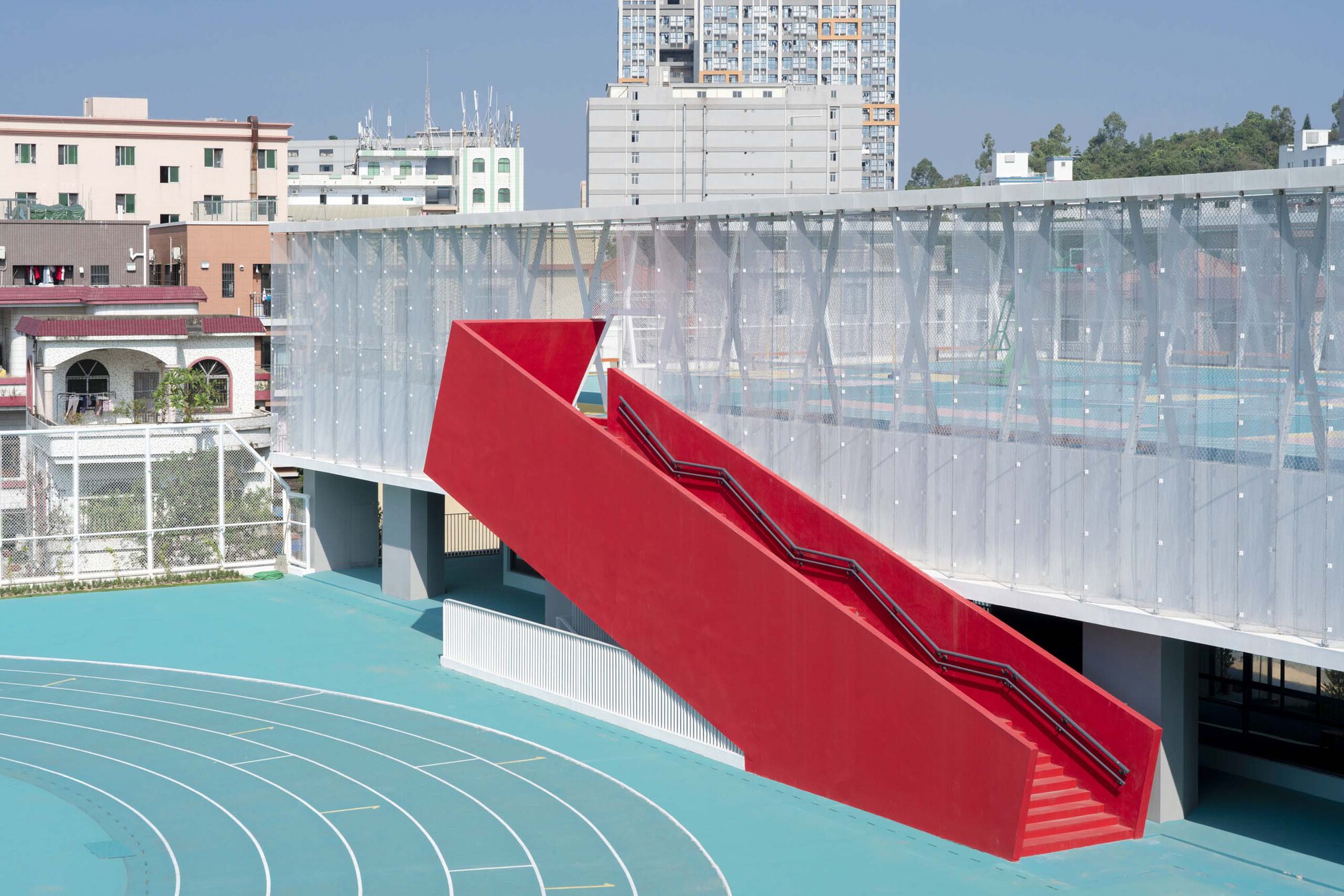 Red sculptural staircase with angled treads rising above the turquoise running track and translucent facade panels