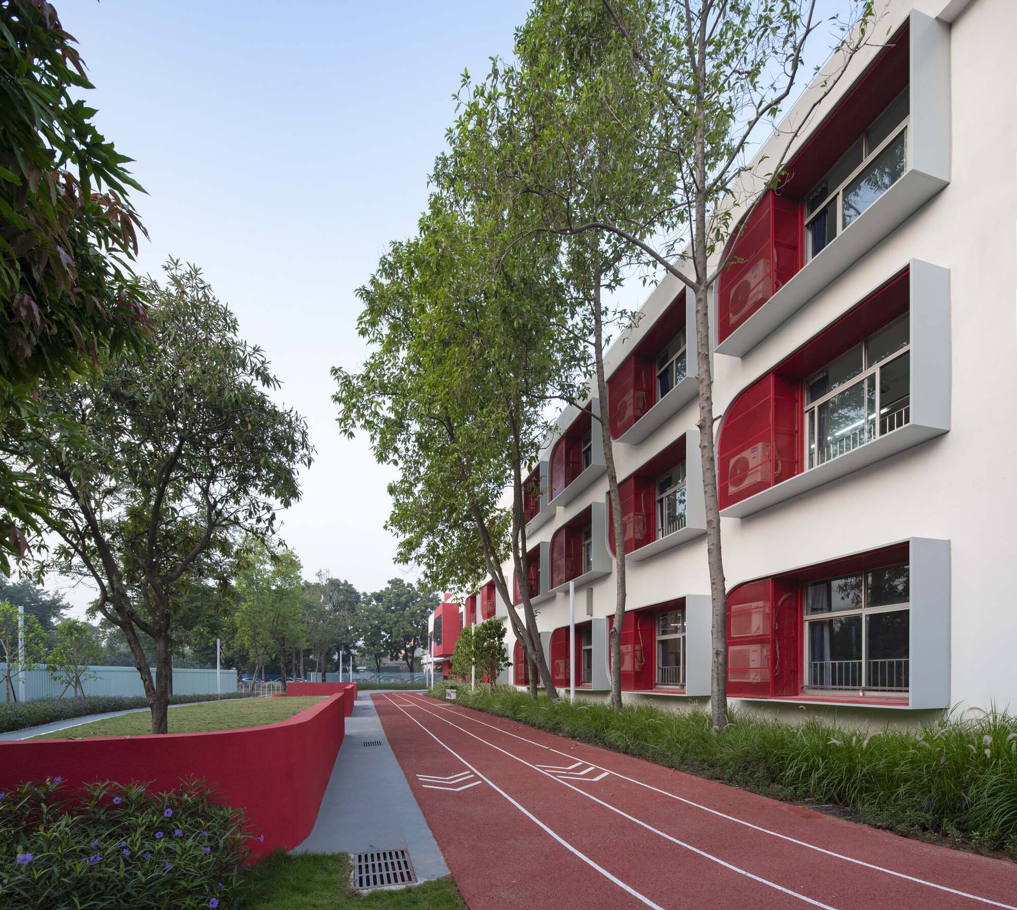 White facade with red panel openings facing a red running track lined with birch trees