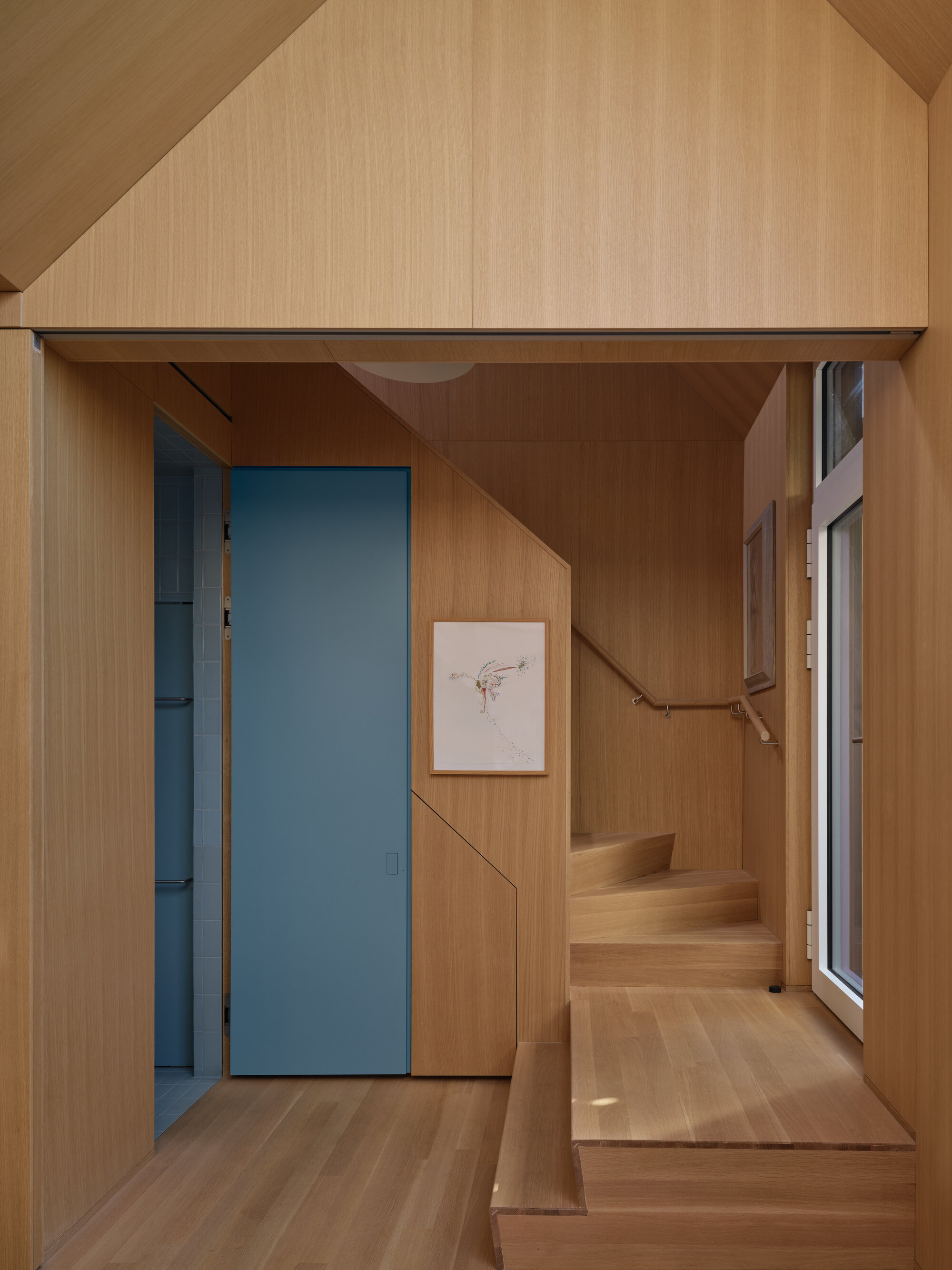 Timber-lined stairwell with flush blue door, built-in seating, and handrail following the ascent