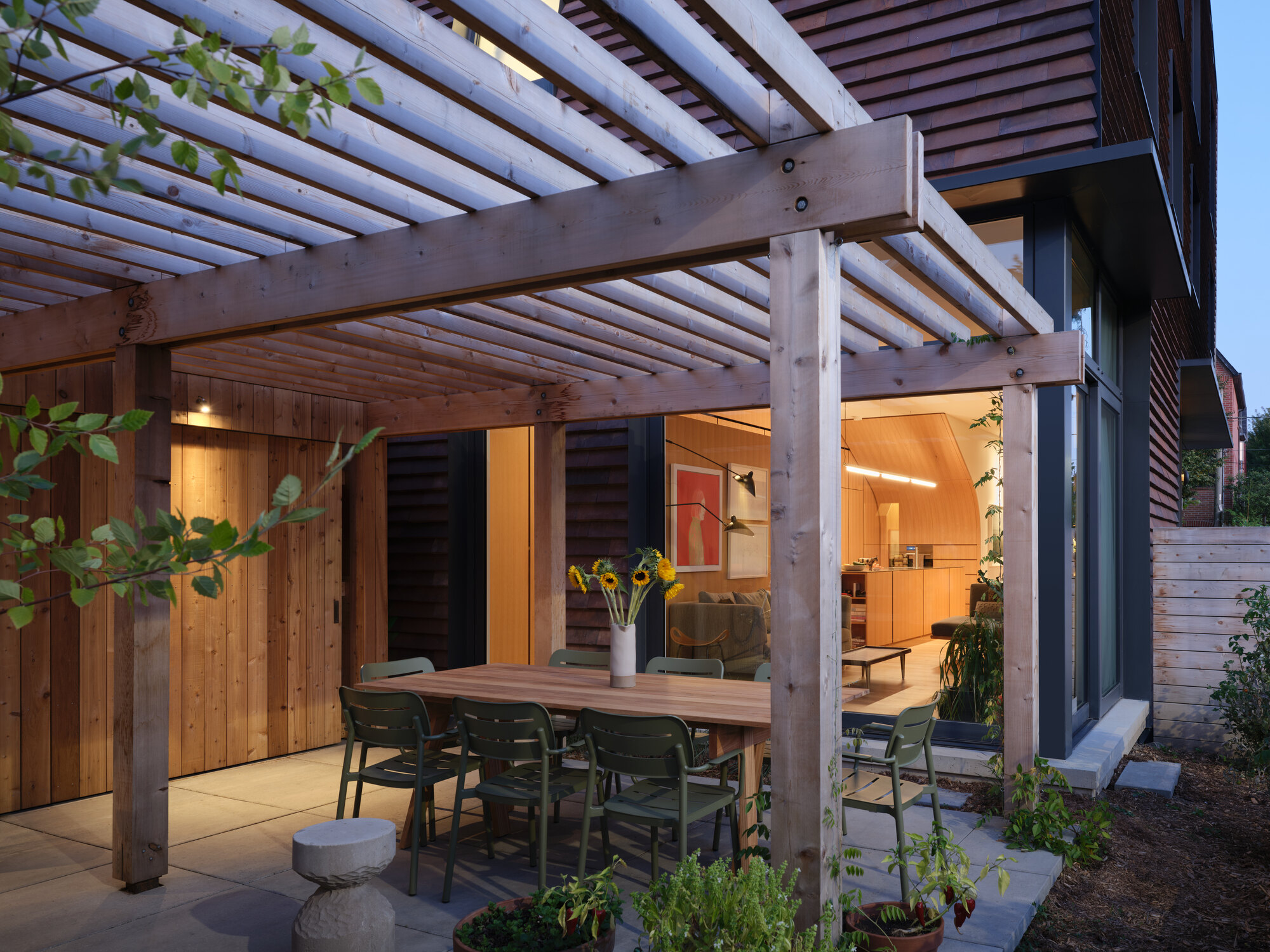 Outdoor dining area under timber trellis structure with potted plants and warm interior lighting