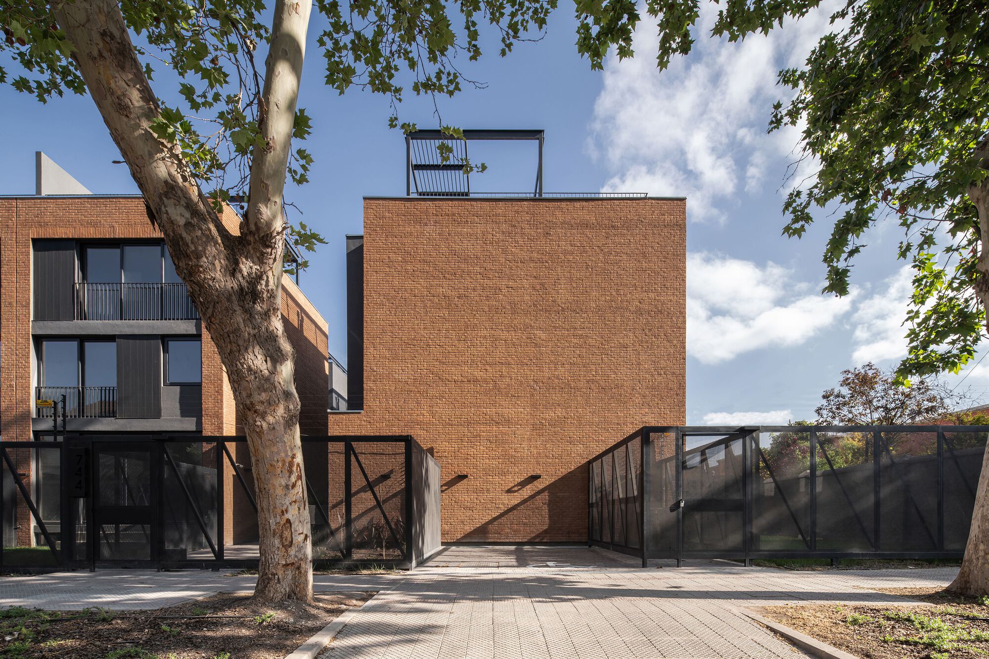 Brick volume with diagonal steel bracing and ramped entrance under afternoon clouds and tree branches