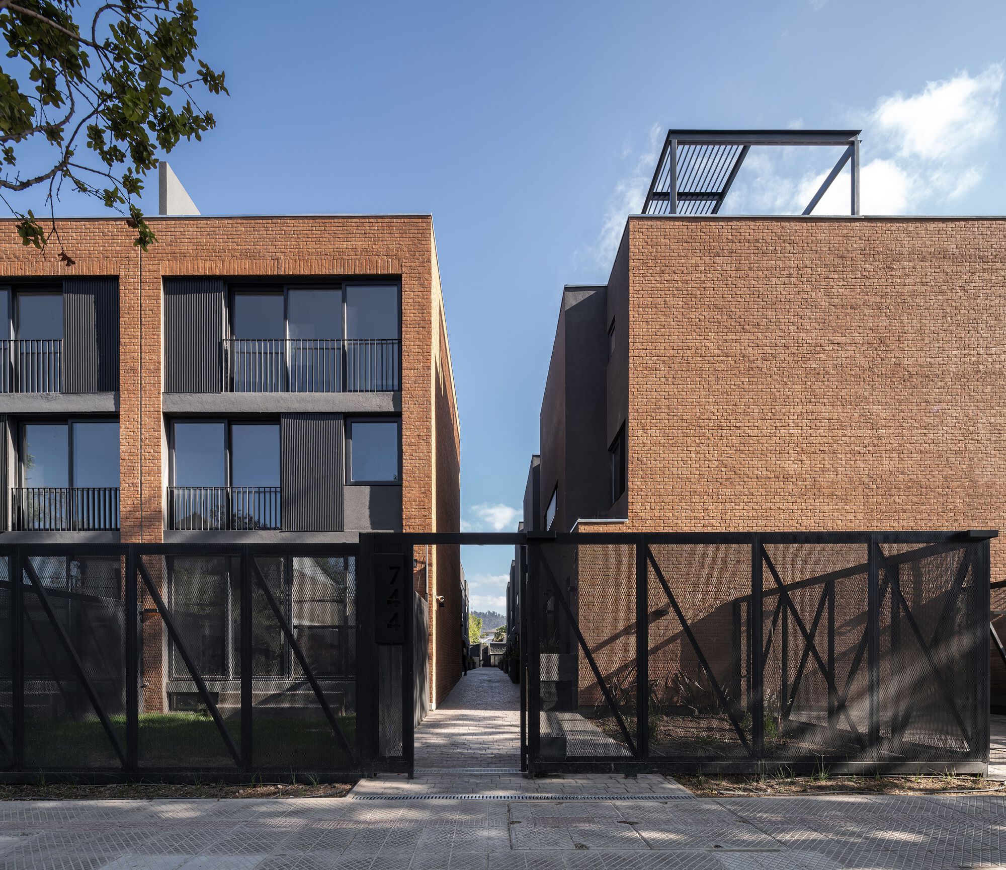 Passageway between two brick volumes with metal rooftop pergola visible against blue sky and clouds
