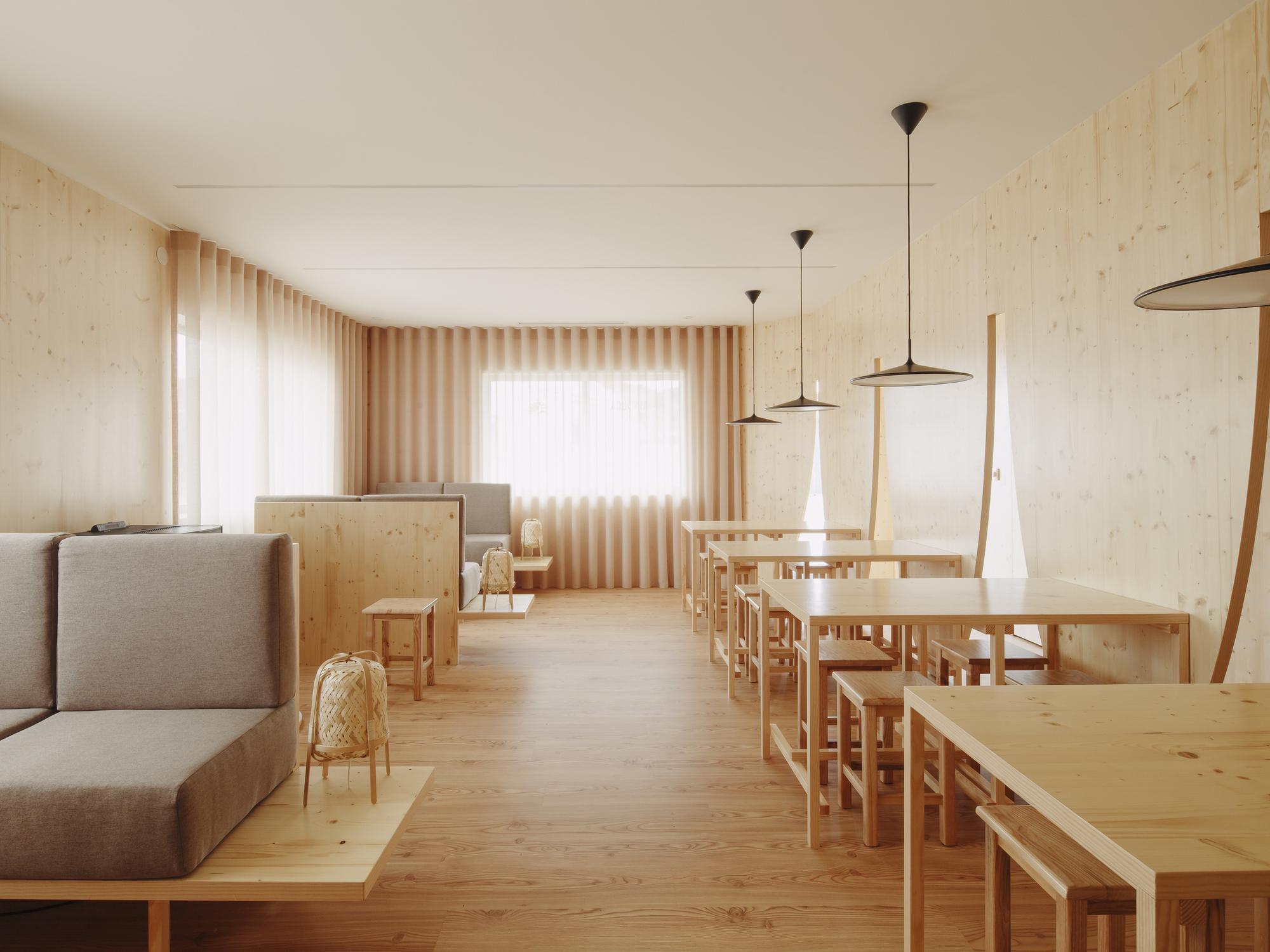 Dining hall with pale timber walls and rows of low tables with pendant lights filtering through sheer curtains
