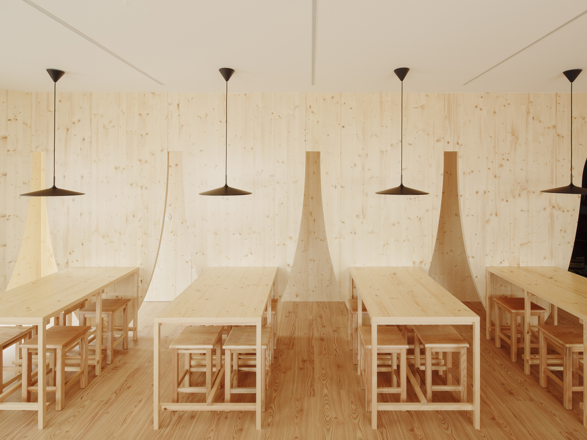 Angled timber partitions creating nooks for dining tables beneath flat pendant lights in a plywood-paneled room
