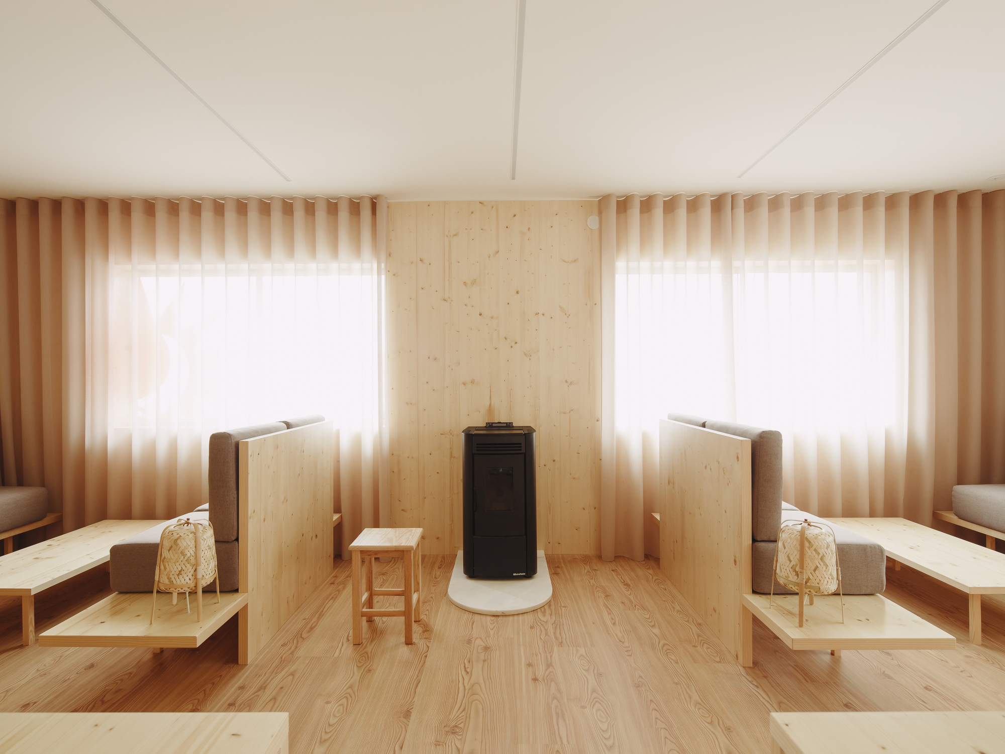 Central wood stove with white base flanked by windows with sheer curtains and timber privacy screens