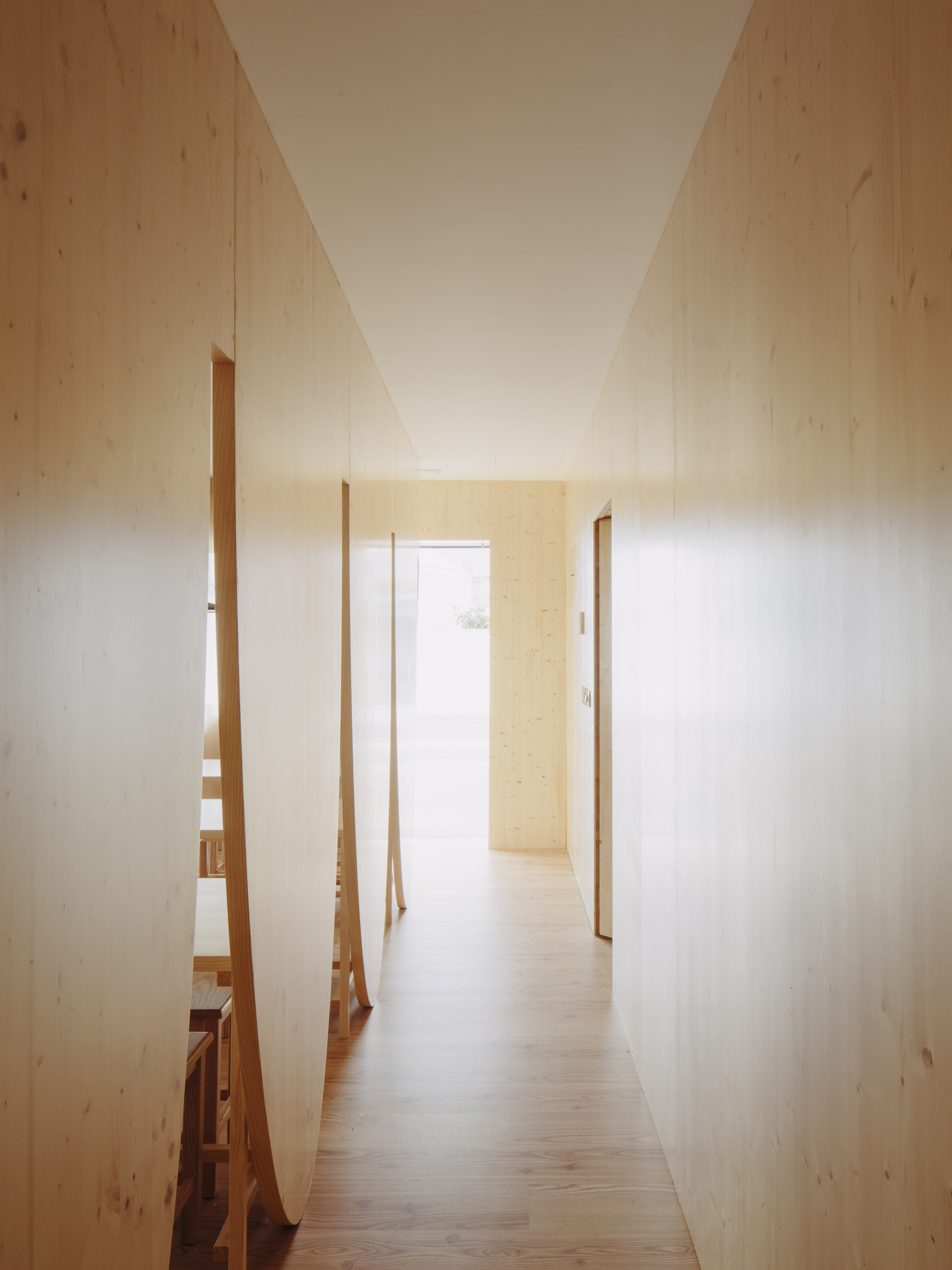 Narrow hallway with pale plywood walls and angled timber partitions opening to a sunlit doorway