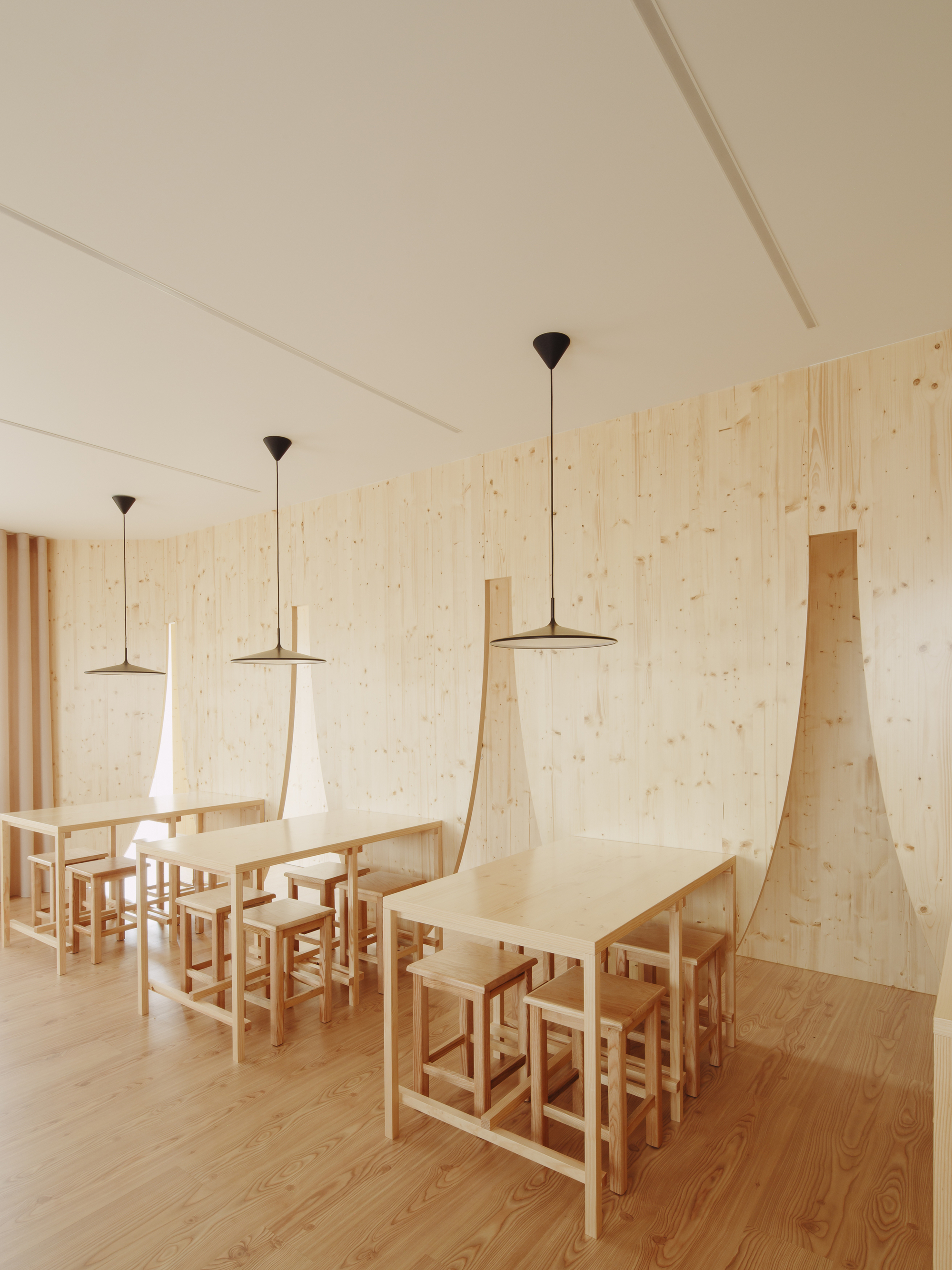 Communal dining tables with pendant lights and angled plywood wall panels beneath a white ceiling