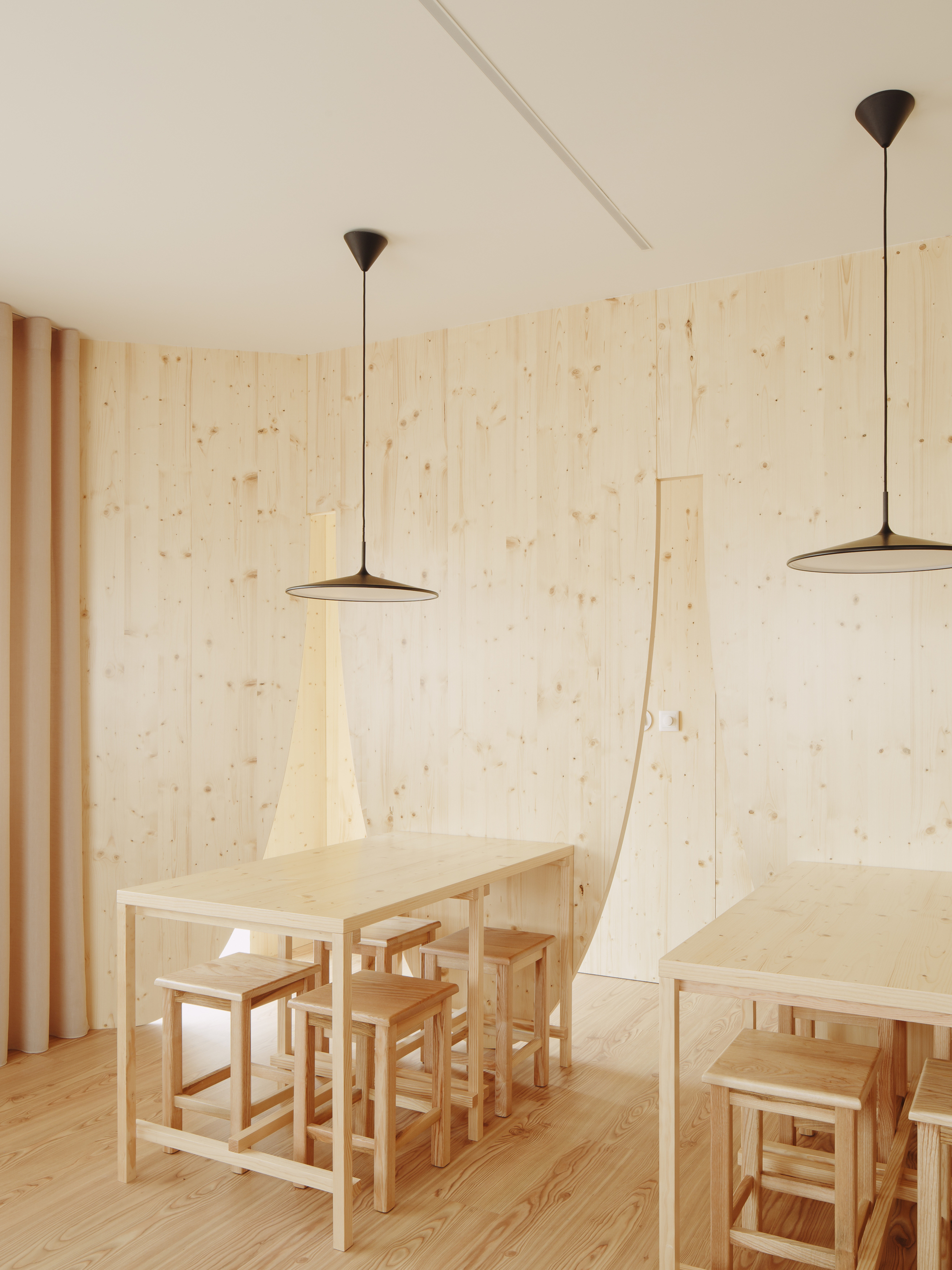 Dining tables with stools and pendant lights against pale plywood walls and vertical drapes