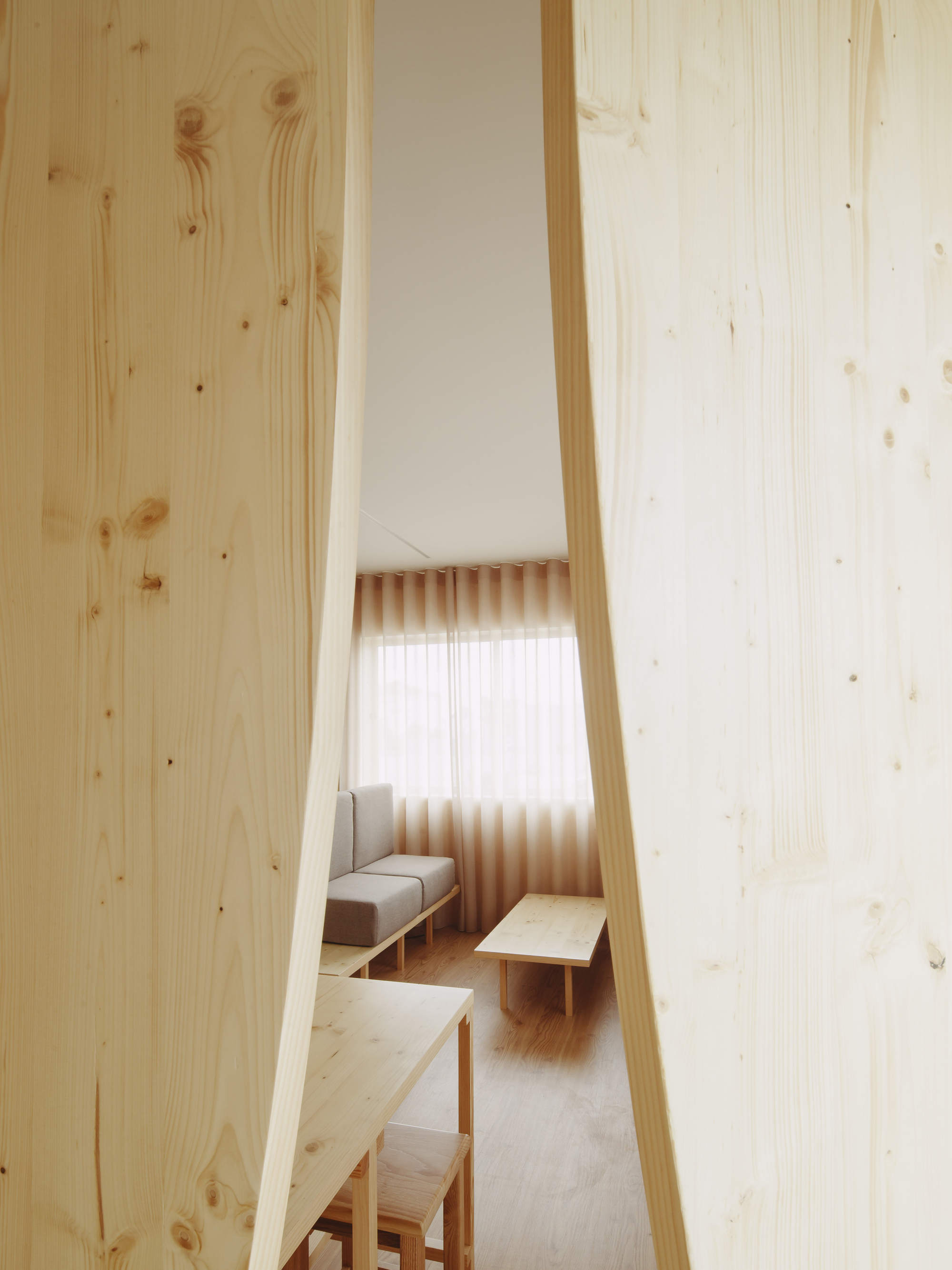 View through a narrow vertical opening in the wood partition toward a seating area with pleated curtain