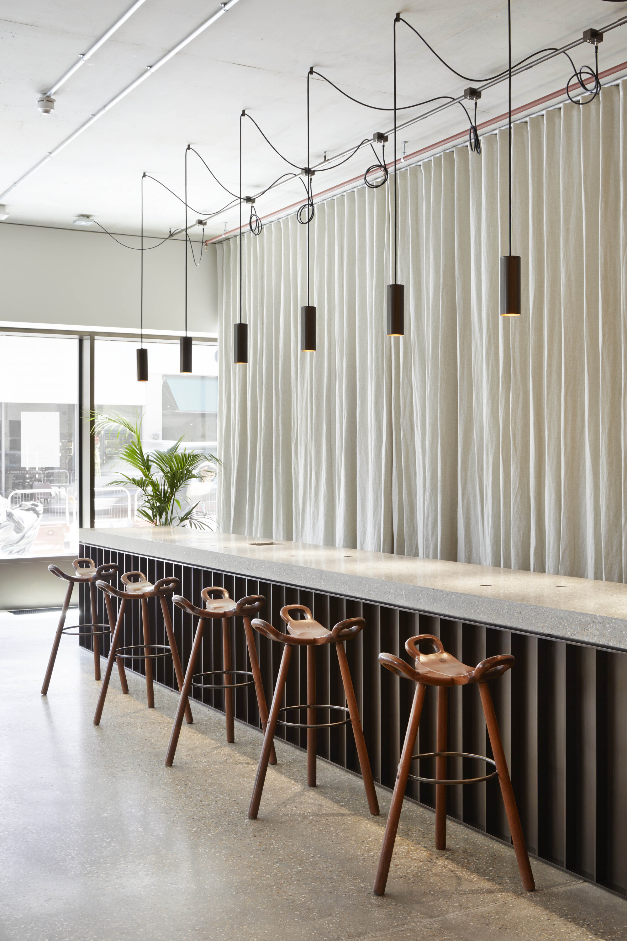 Long bar counter with timber stools below cylindrical pendant lights suspended from exposed conduit