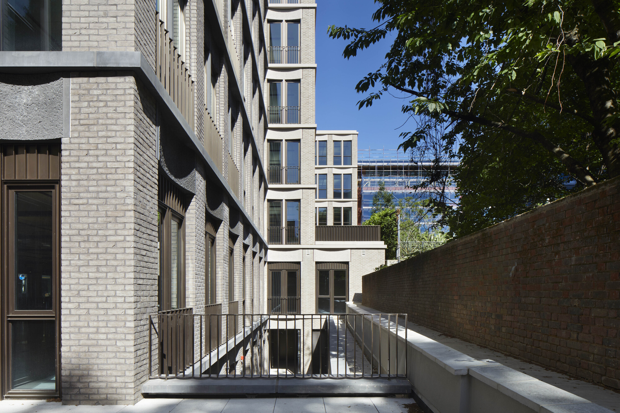 Courtyard view showing brick walls and metal railings framing a sunlit passage with tree foliage