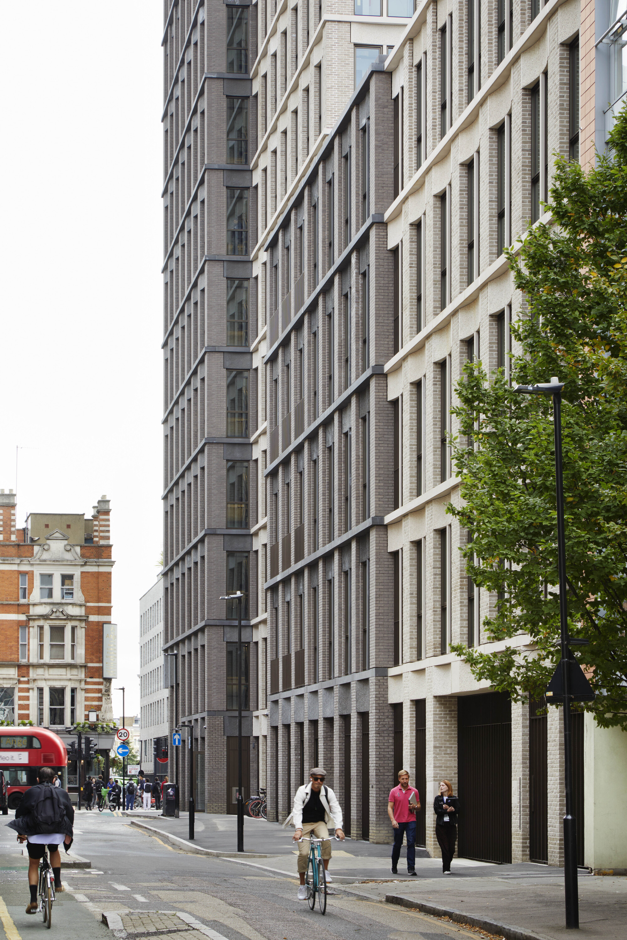 Street view of dark brick facade with vertical window recesses and cyclists passing below