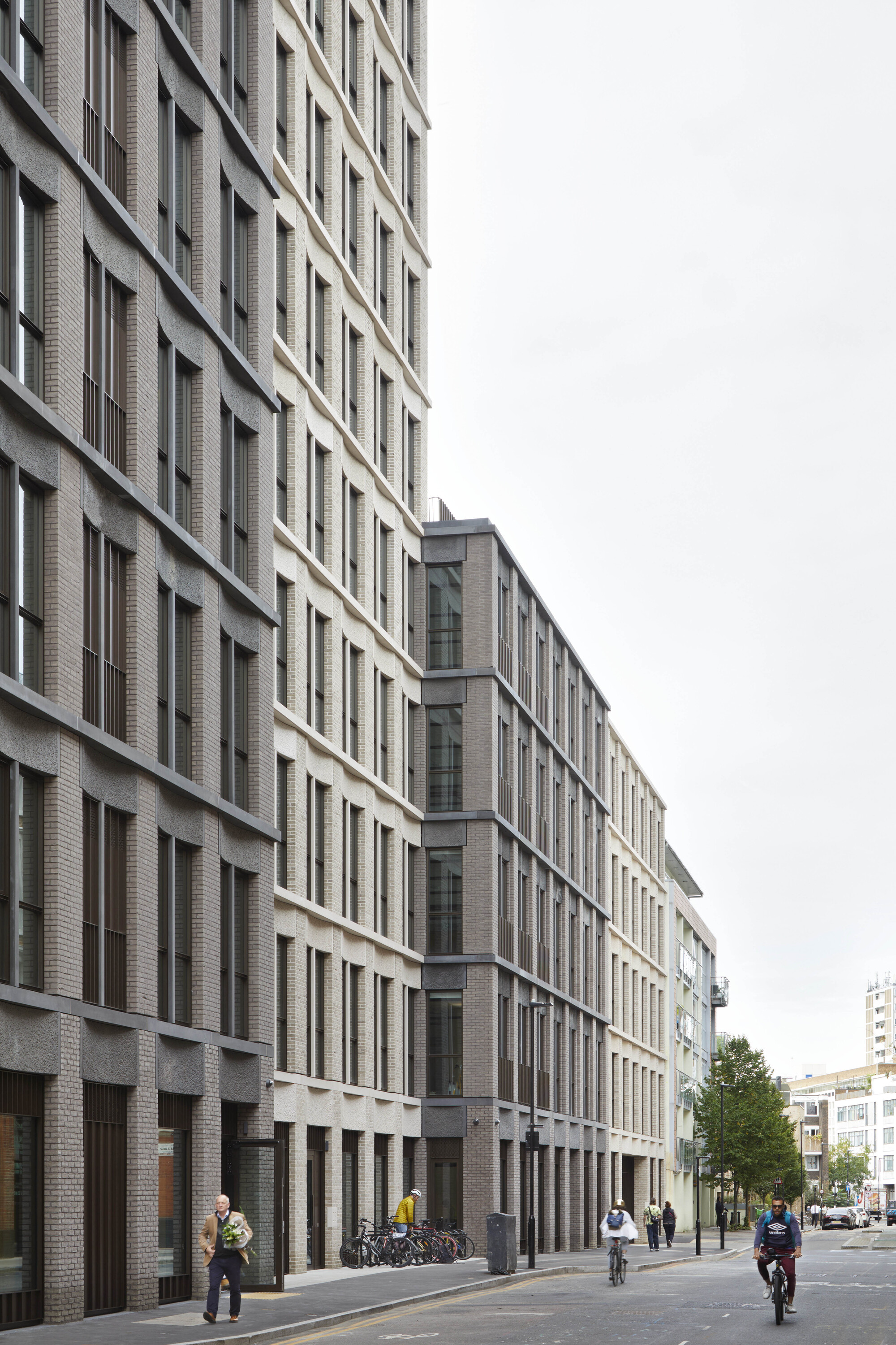 Grey brick facade along a street with pedestrians and cyclists under overcast sky