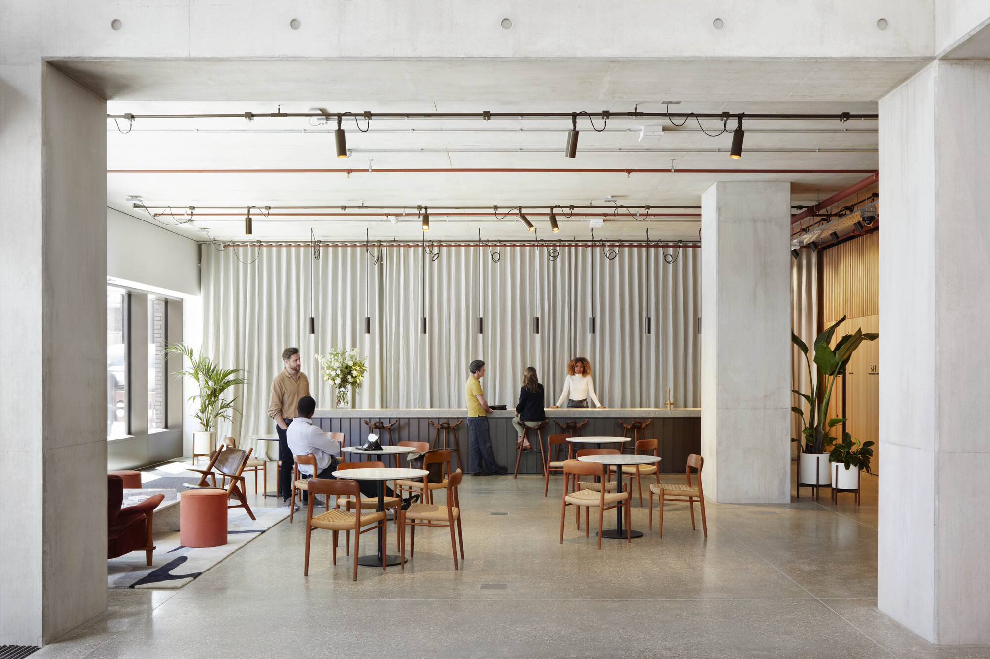 Open lounge area with exposed concrete beams, timber furniture and people gathering beneath pendant lighting
