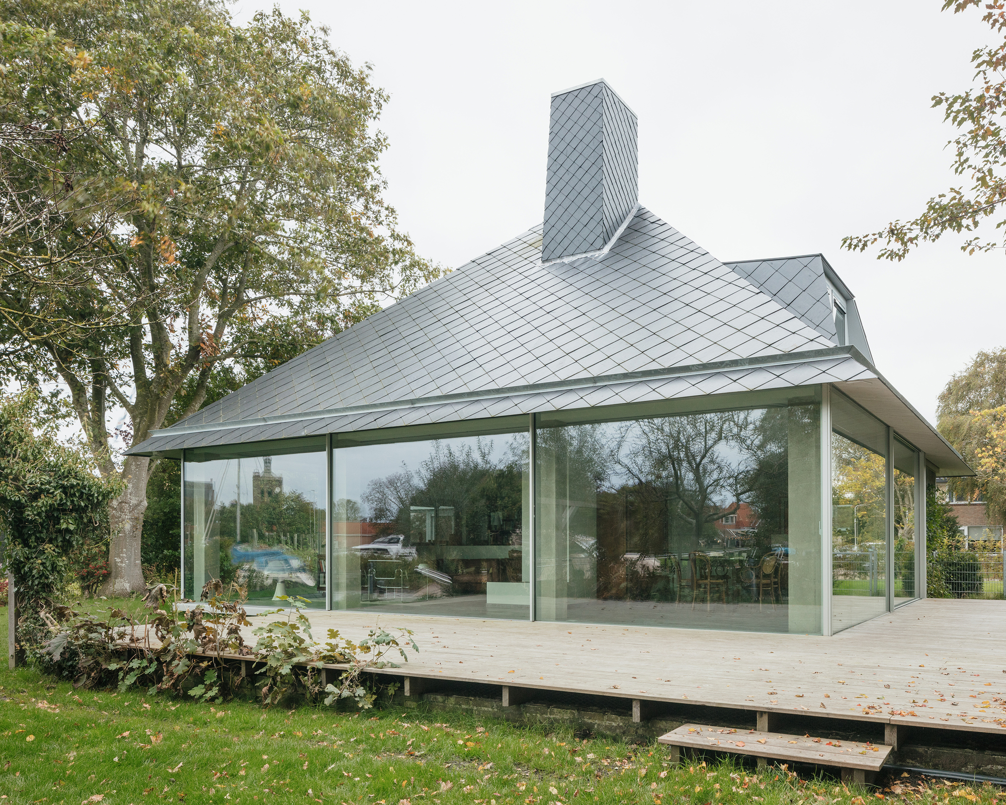 Glass-enclosed pavilion with sliding panels raised on a timber deck surrounded by lawn