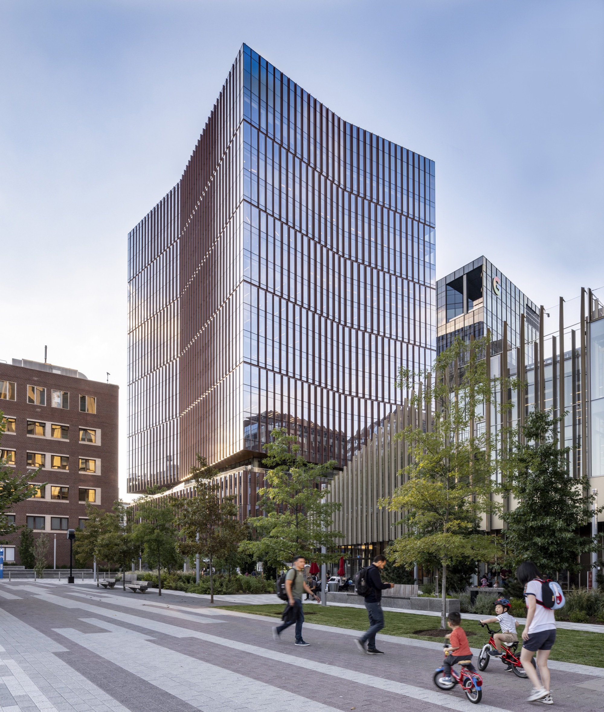 Vertical glass and fin facade with families on bicycles passing through the landscaped plaza at dusk