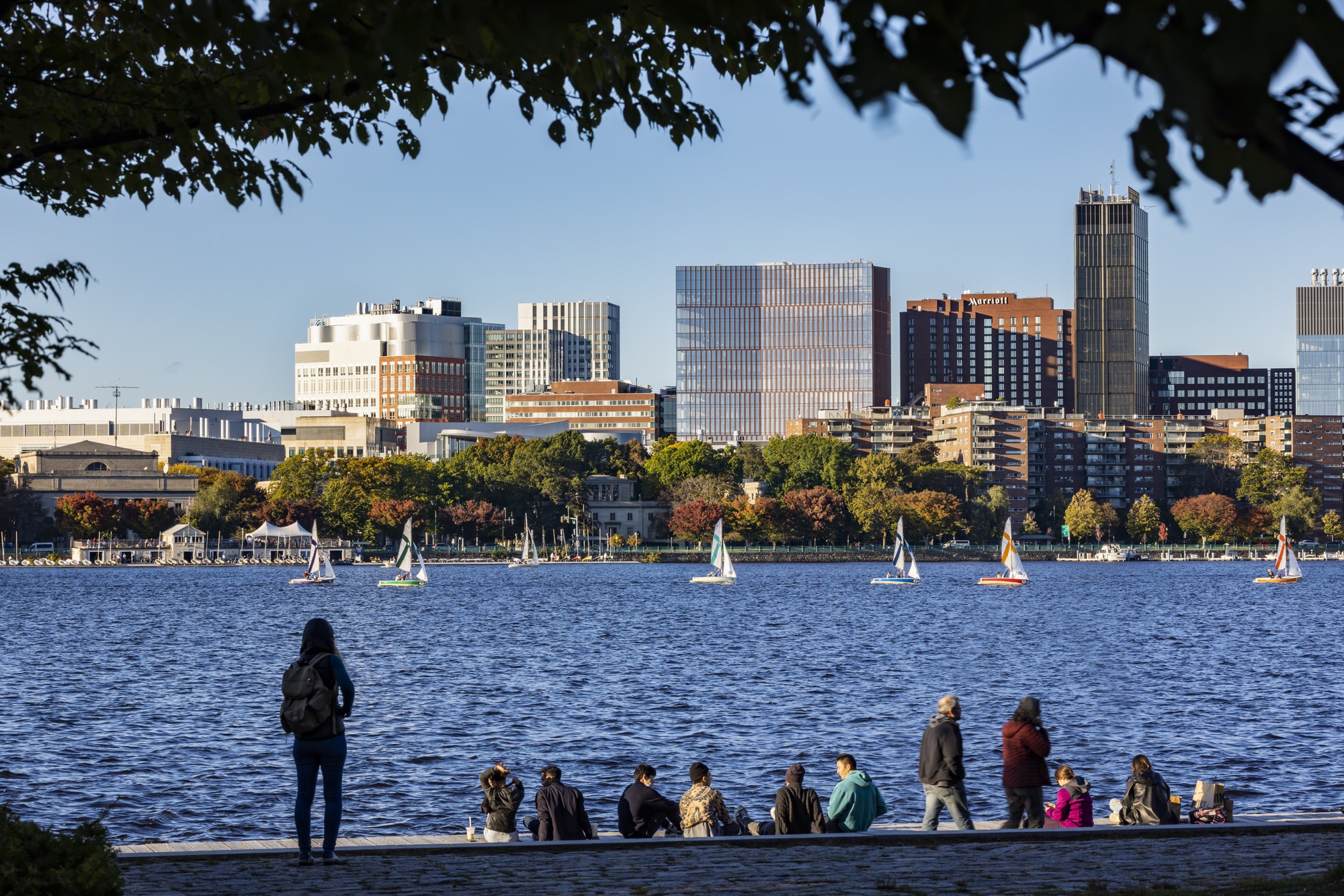 Skyline view from across the river with sailboats on the water and people gathered along the shore