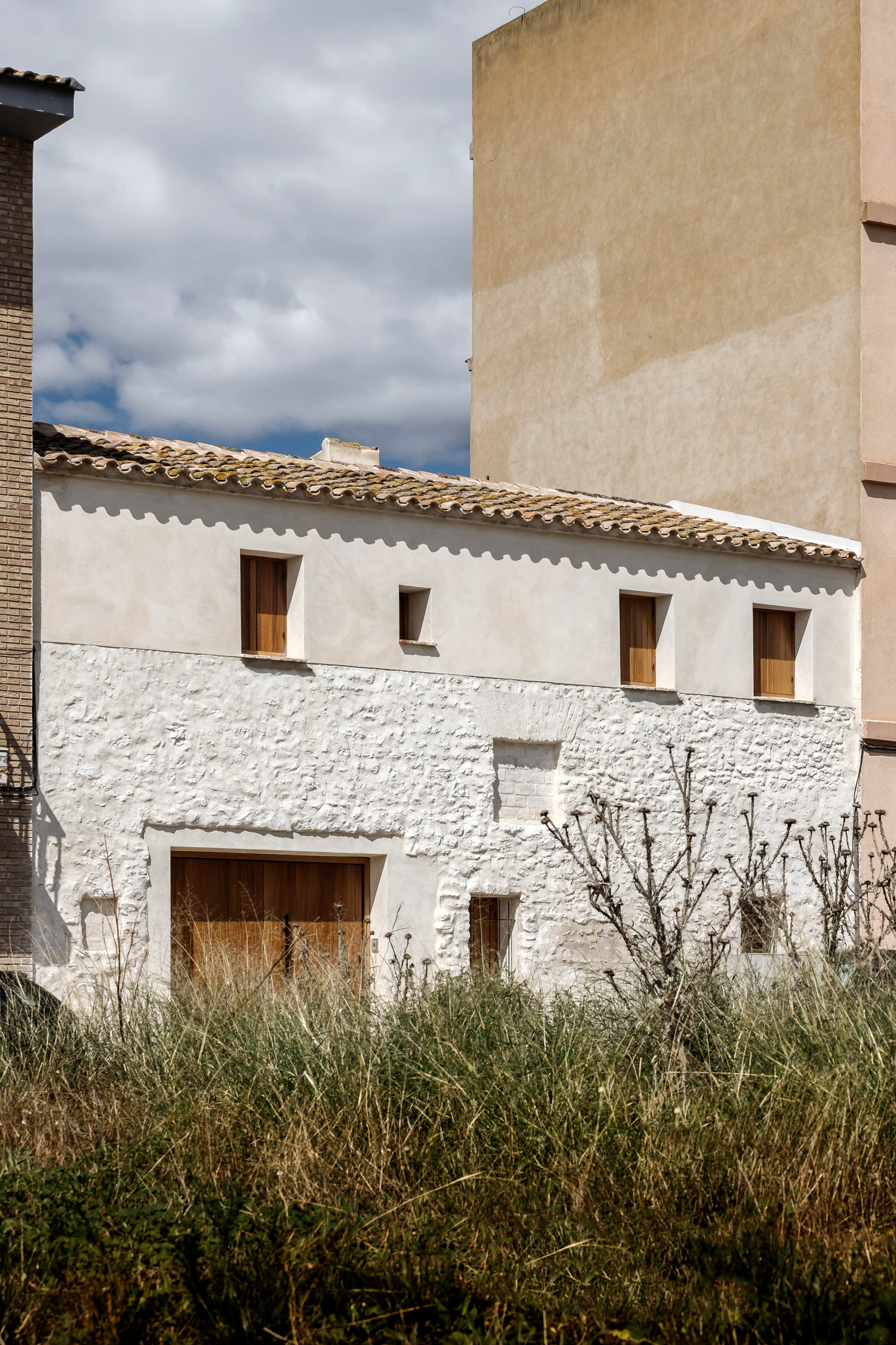 White rendered two-story facade with timber openings and terra-cotta tiled roof under cloudy sky