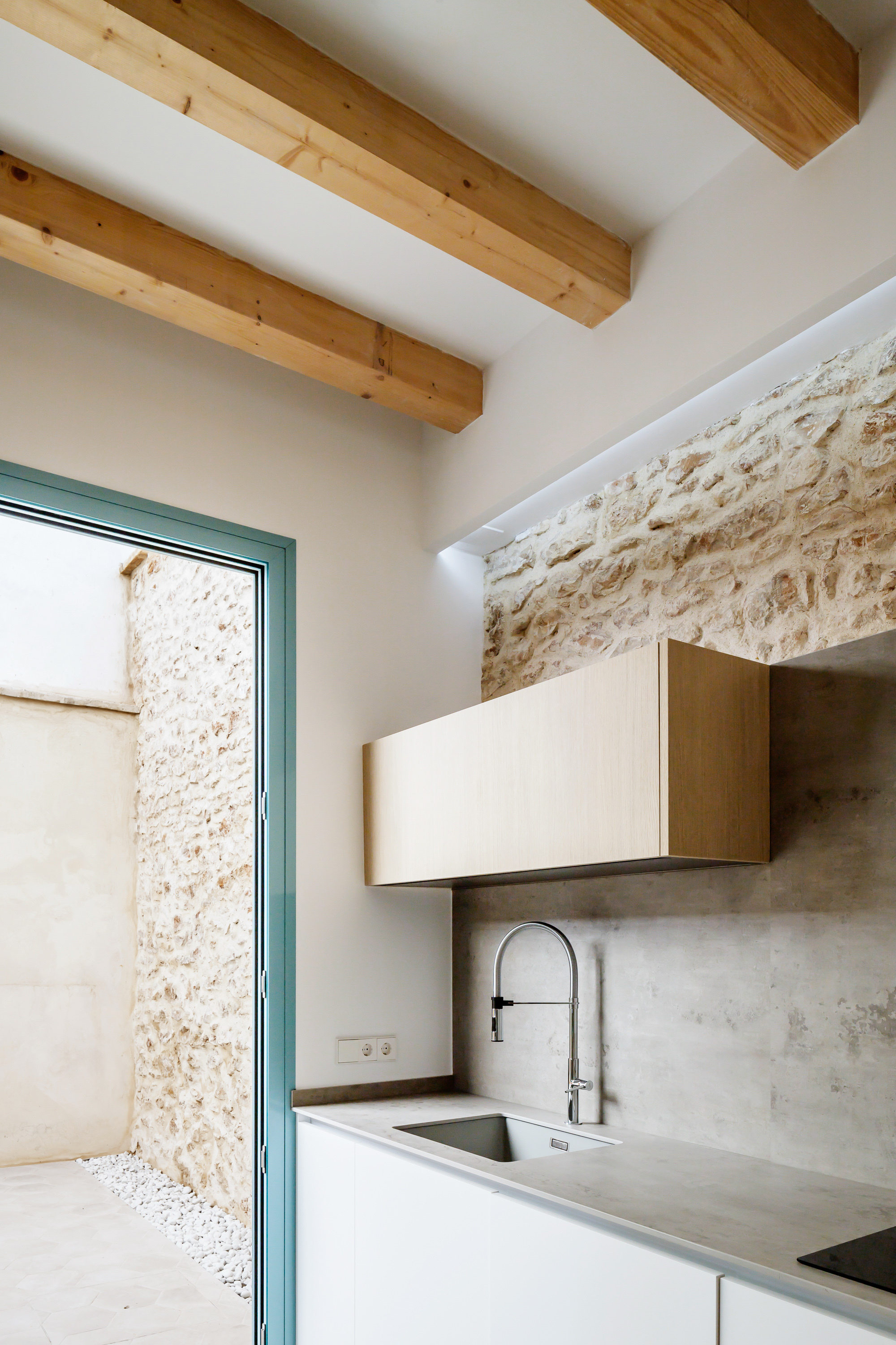 Kitchen with white cabinetry and concrete countertop beneath exposed timber ceiling beams and stone wall