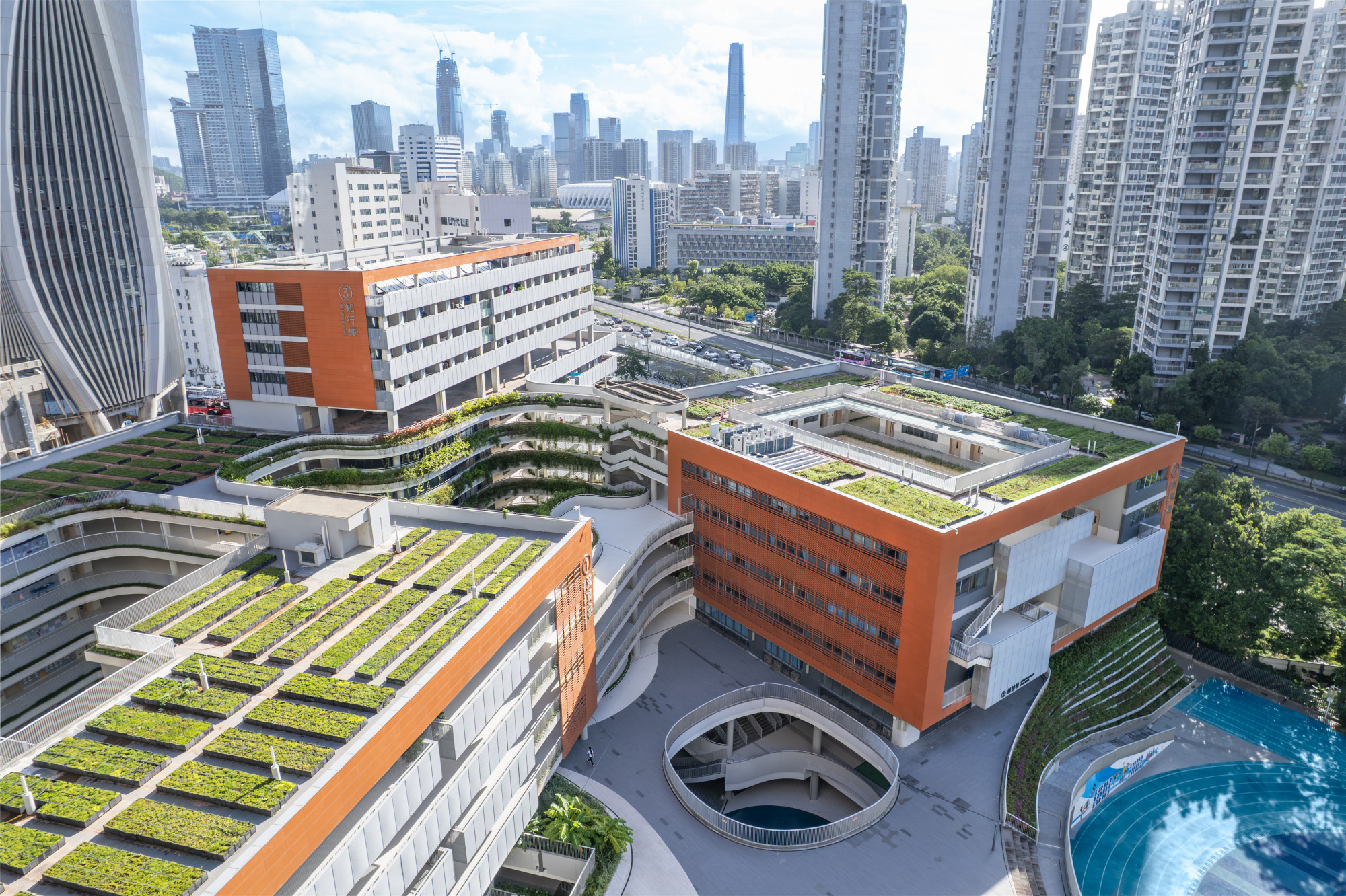 Aerial view of terraced green roofs and orange-clad volumes with a skyline backdrop under clear blue sky