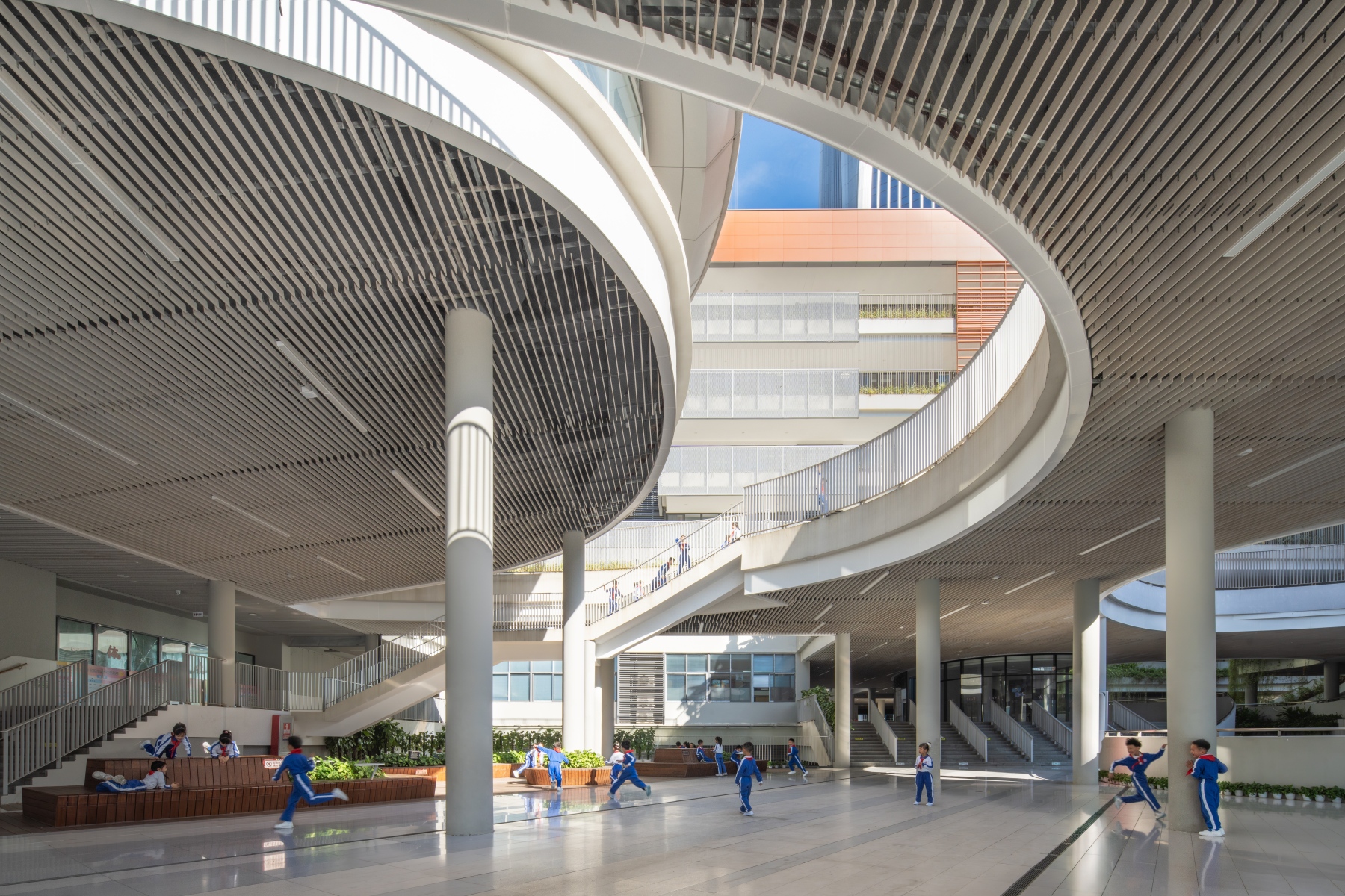 Covered ground-level plaza with white columns, wood-slatted ceiling, and a sweeping curved ramp with students in blue uniforms