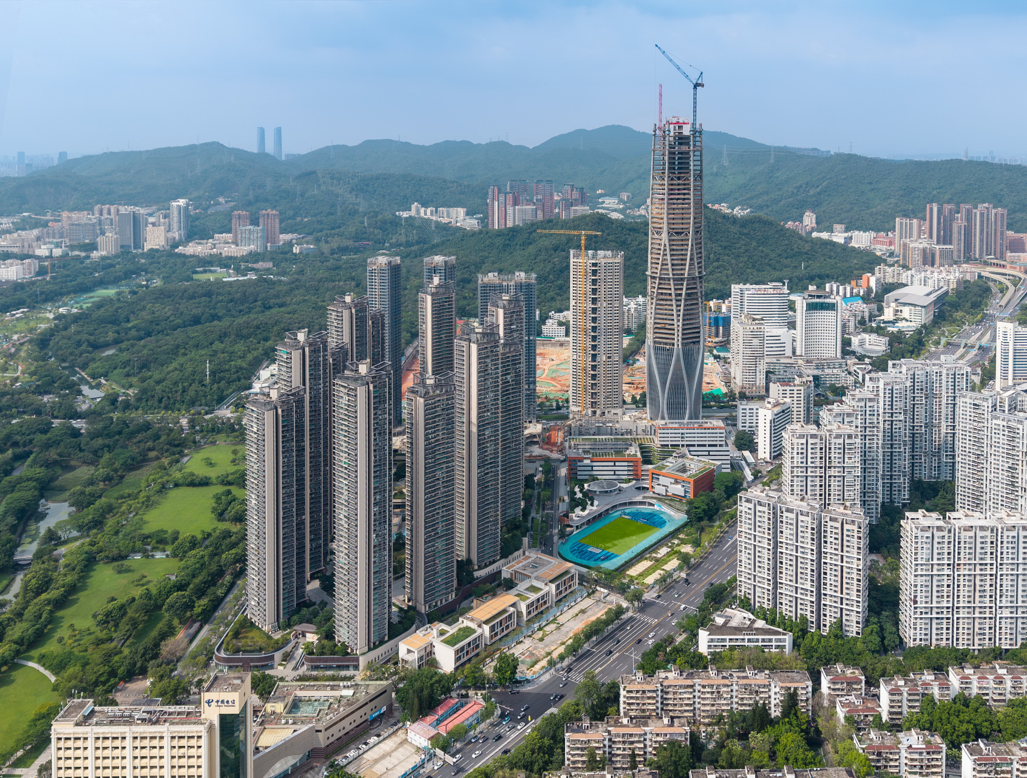 Aerial view of paired residential towers rising above a landscaped podium with sports fields and forested hills beyond