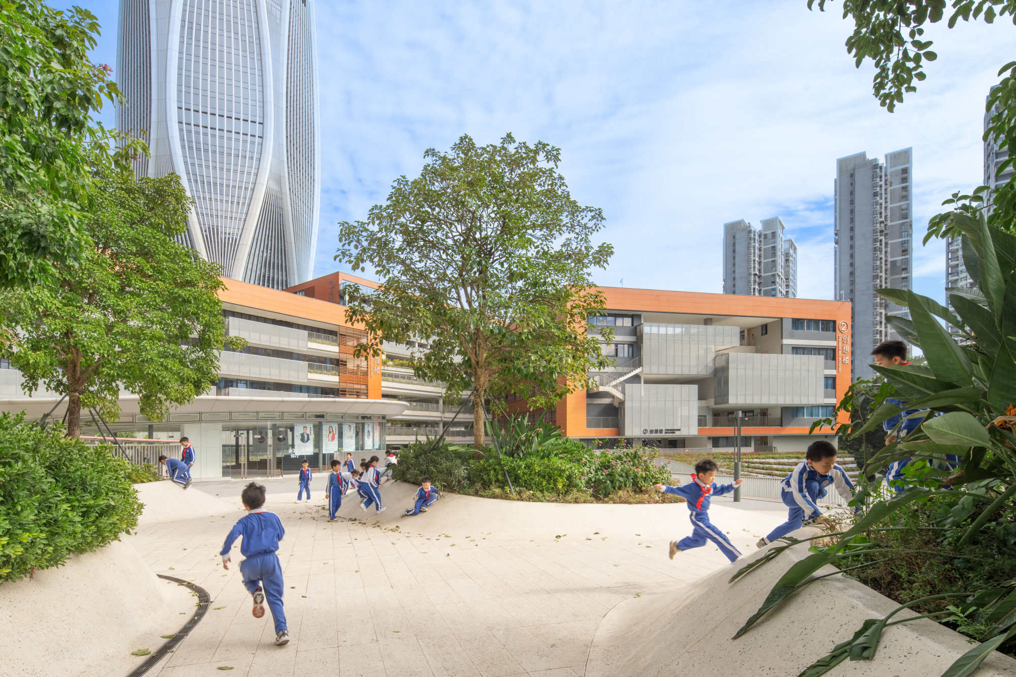 View of the orange-banded school building across a sand play area with children running and trees framing the scene