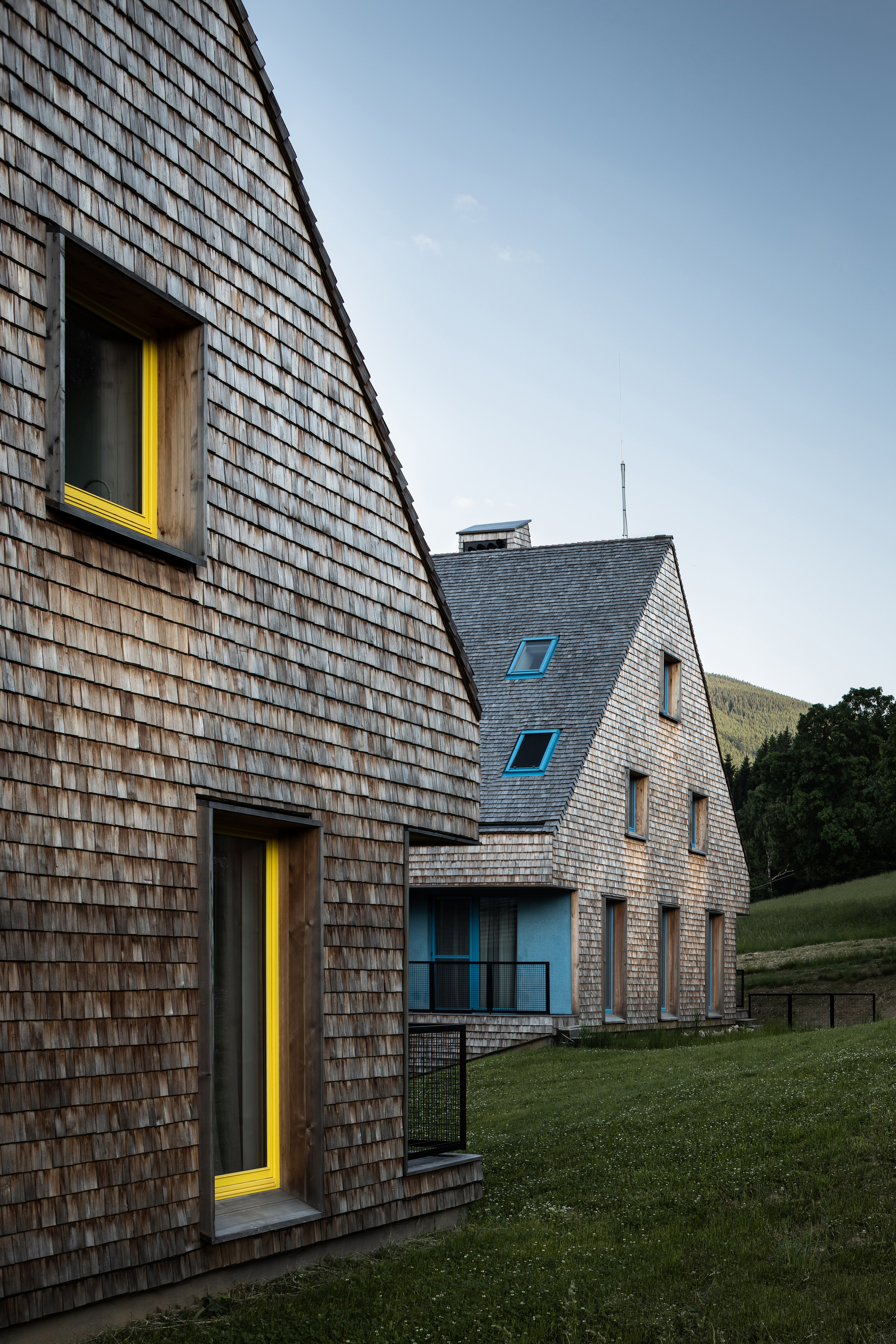Steeply pitched shingled facade with yellow door frames at dusk on sloping grass terrain