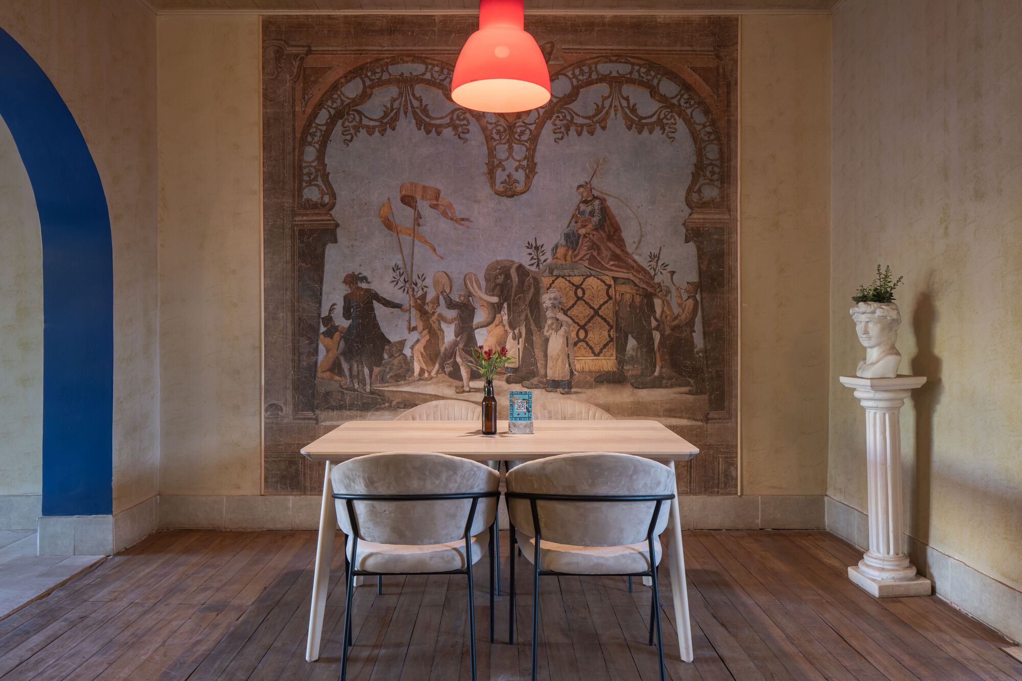 Dining alcove with faded fresco wall mural and red pendant light above timber floor