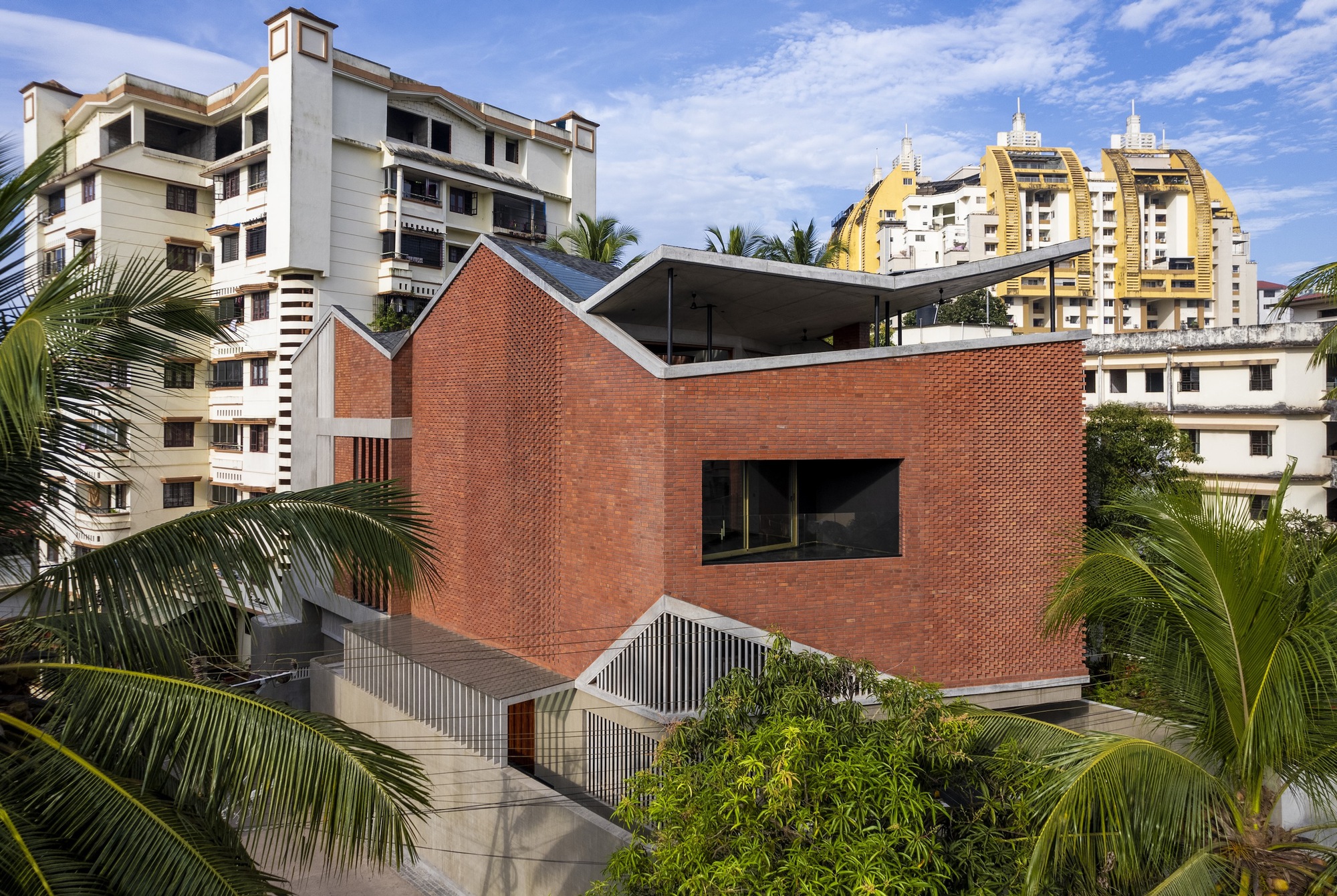 Elevated view of the red brick facade with perforated screens and folded roof amid palm trees and residential towers