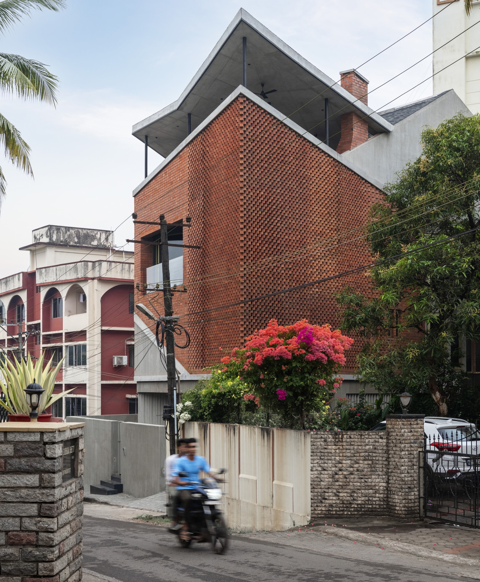 Street view showing the angled brick facade with a motorbike passing and flowering plants at the boundary wall