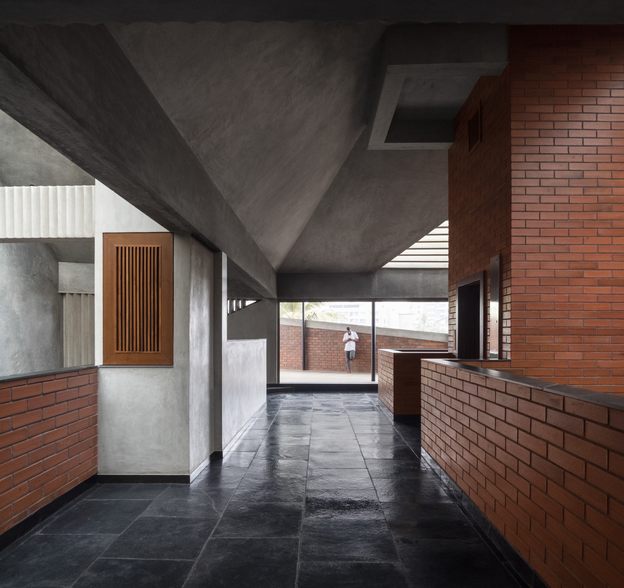 Interior corridor with exposed concrete ceiling and brick half-walls leading to a terrace with a figure walking