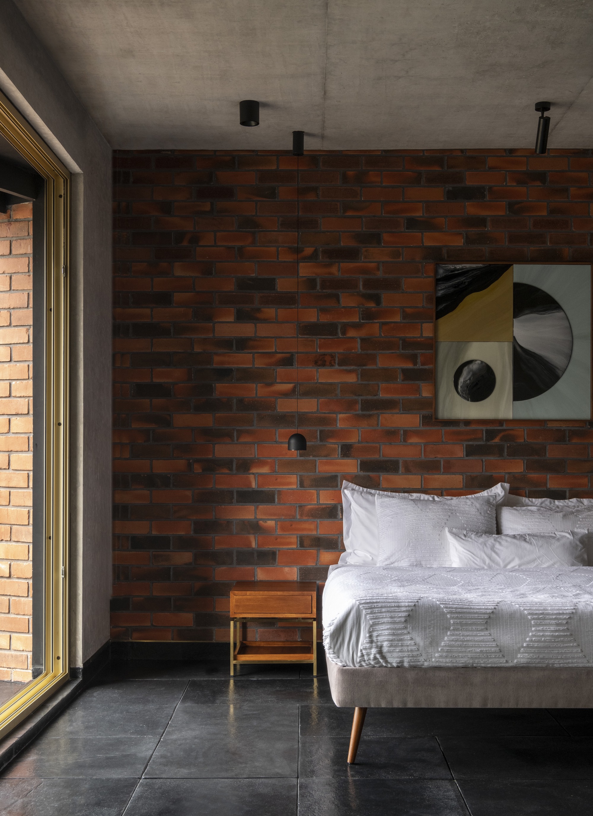 Bedroom with variegated red brick wall, exposed concrete ceiling and dark stone floor beside a glazed opening