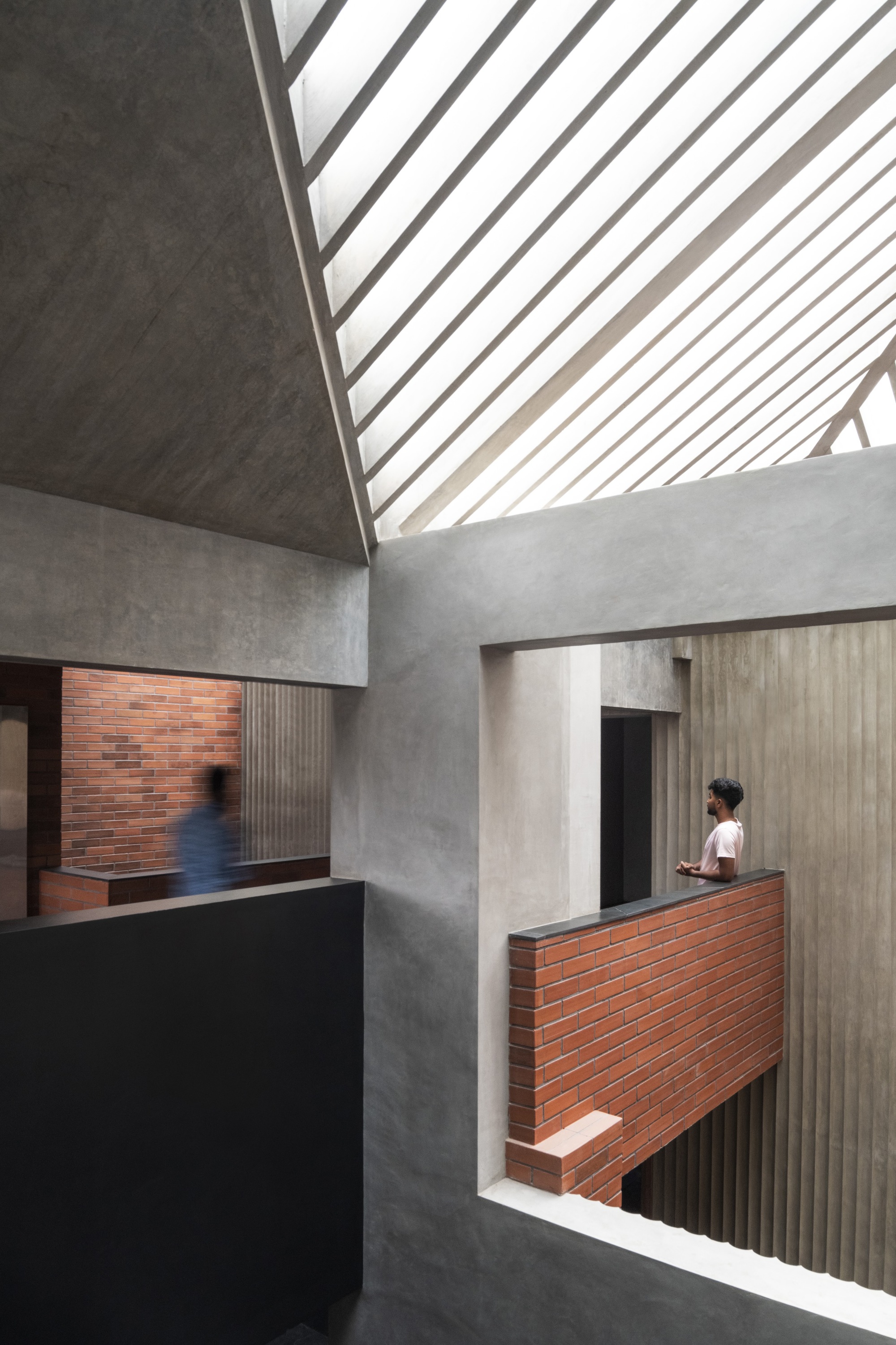 Double-height interior space with slatted skylight casting diagonal shadows across concrete walls and brick balcony where a person stands