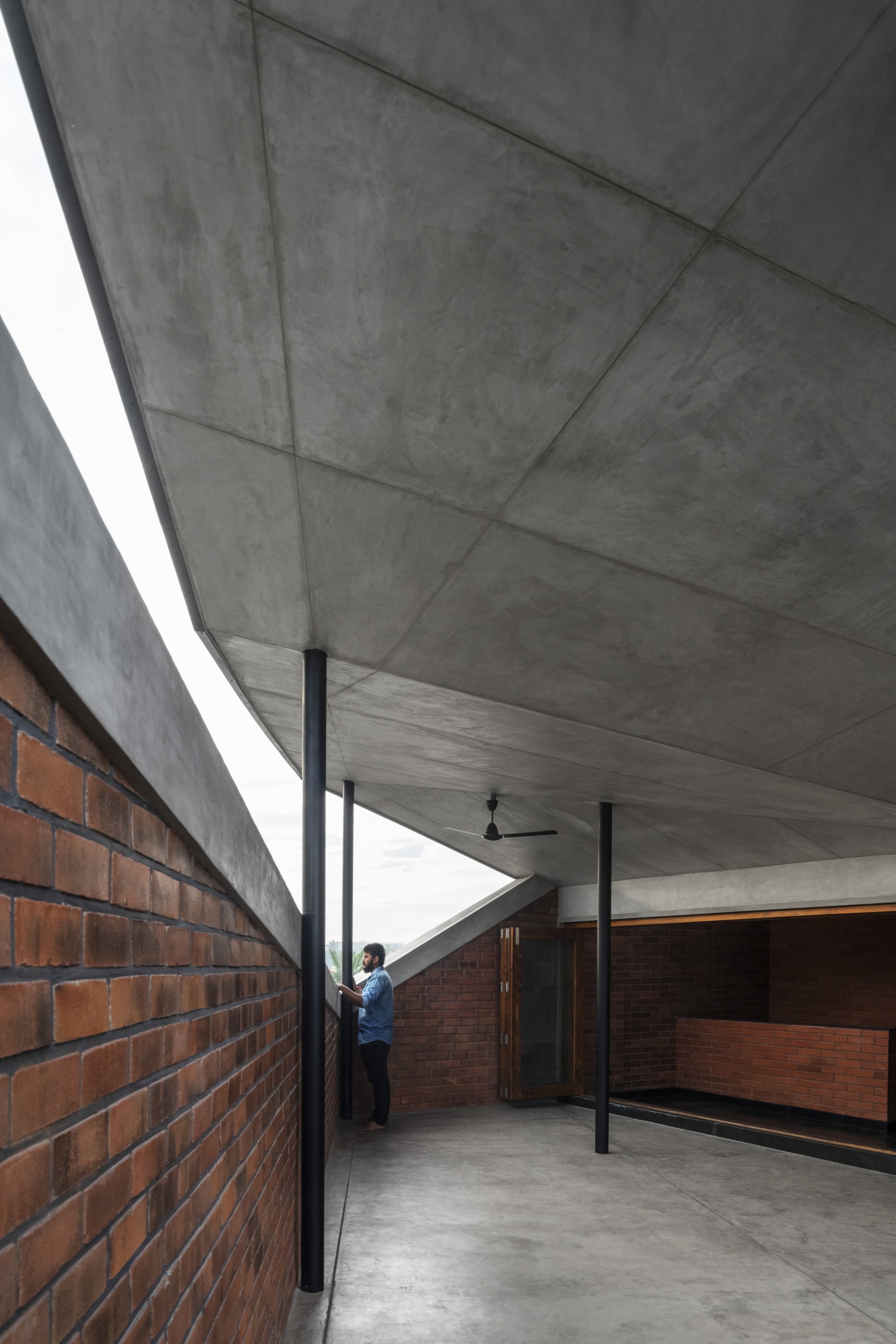 Covered terrace with exposed concrete ceiling, black steel columns, and red brick walls as a person stands in the passage