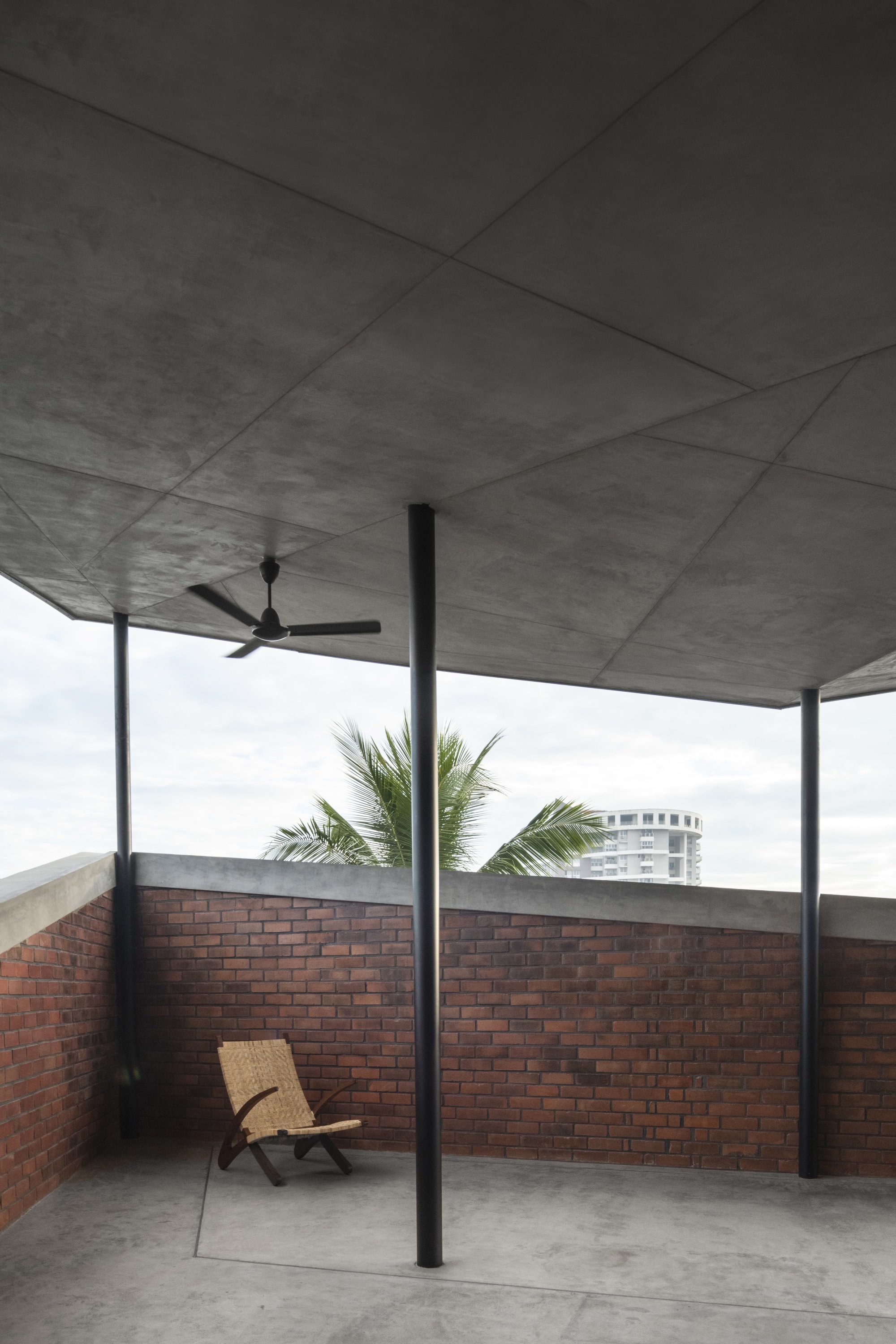 Rooftop terrace with concrete soffit, ceiling fan, steel columns, and brick parapet walls with palm fronds visible beyond