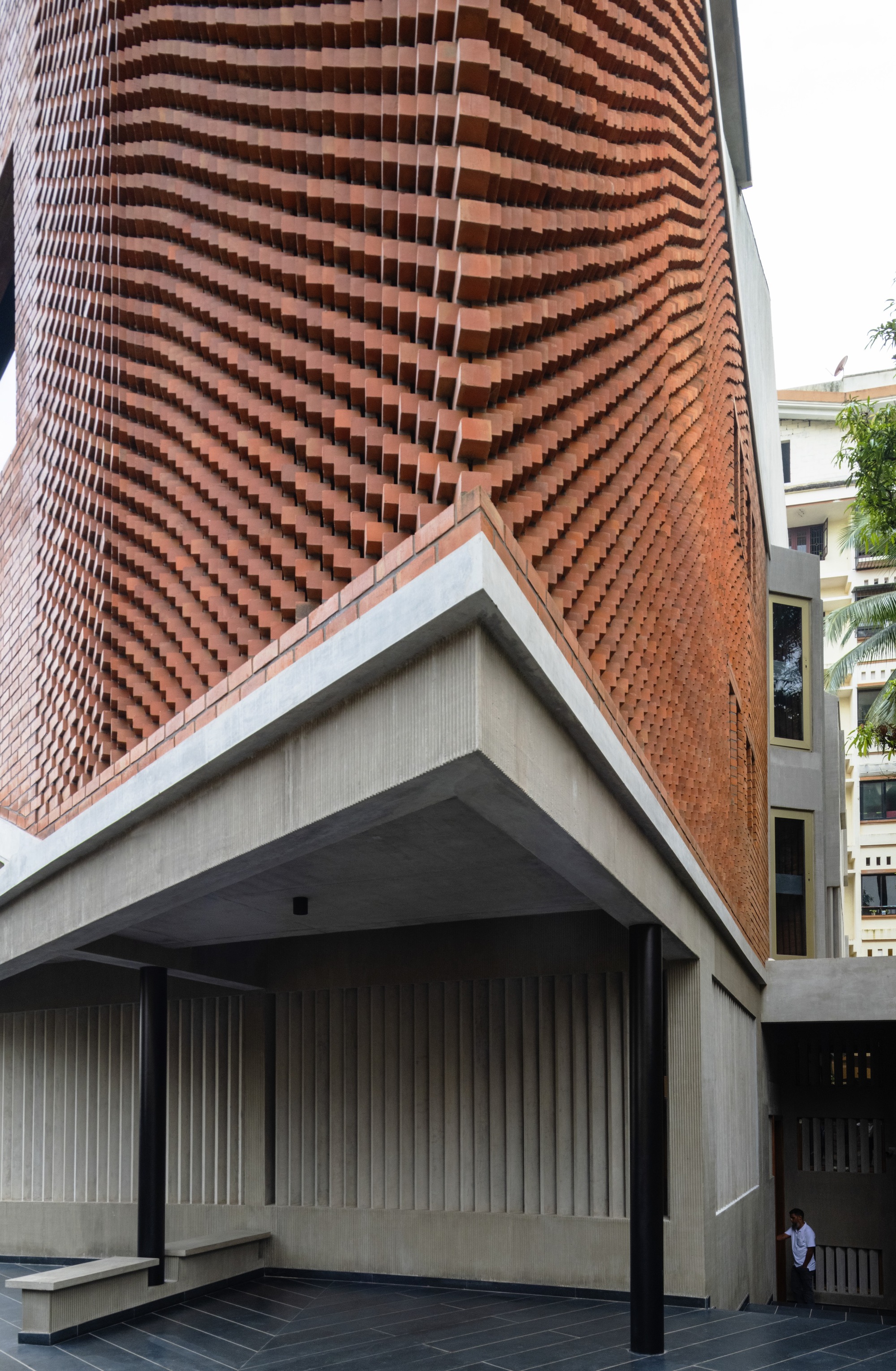 Undulating chequered brick facade meeting angular concrete soffit above a covered entry with vertical timber cladding