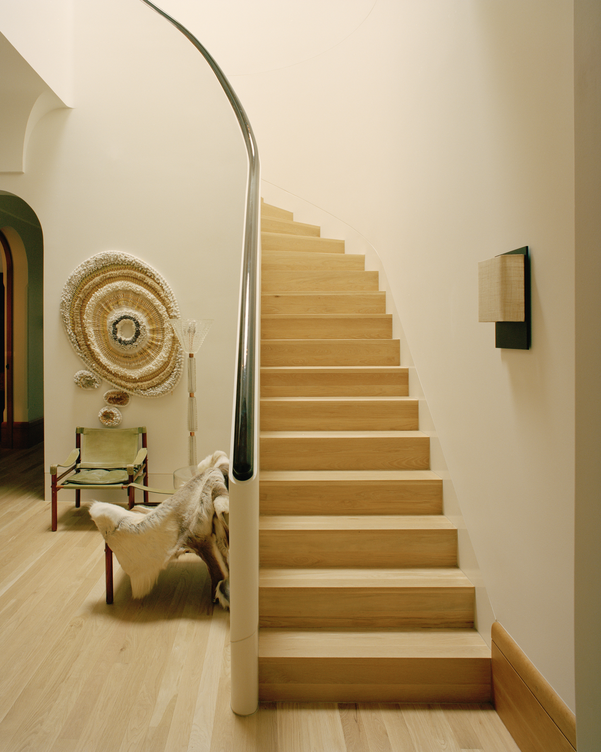 Oak staircase with curved steel handrail ascending beside white walls and a chair with woven wall hanging