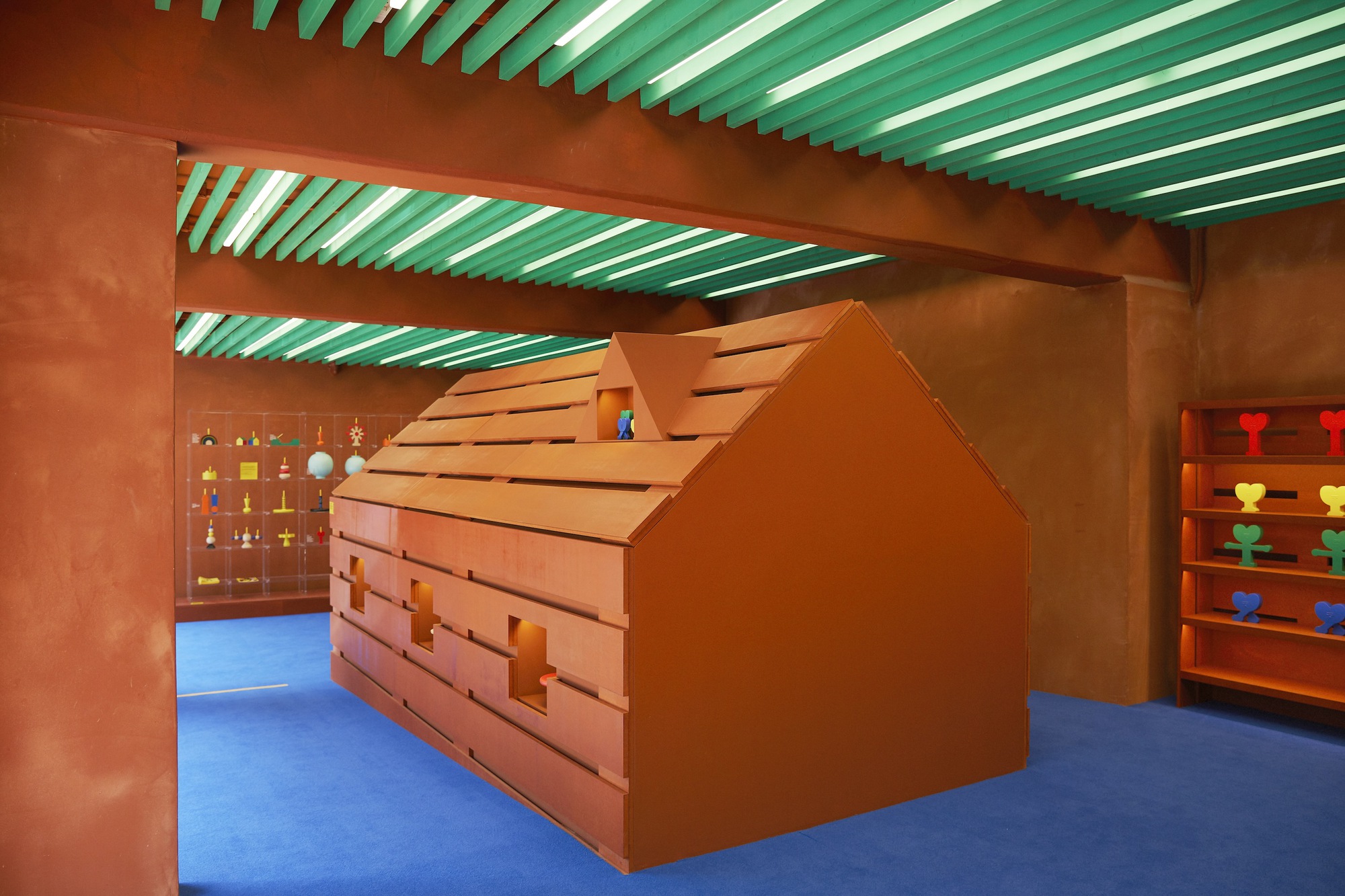 House-shaped timber storage structure with slot openings beneath green slatted ceiling beams and blue rubber flooring