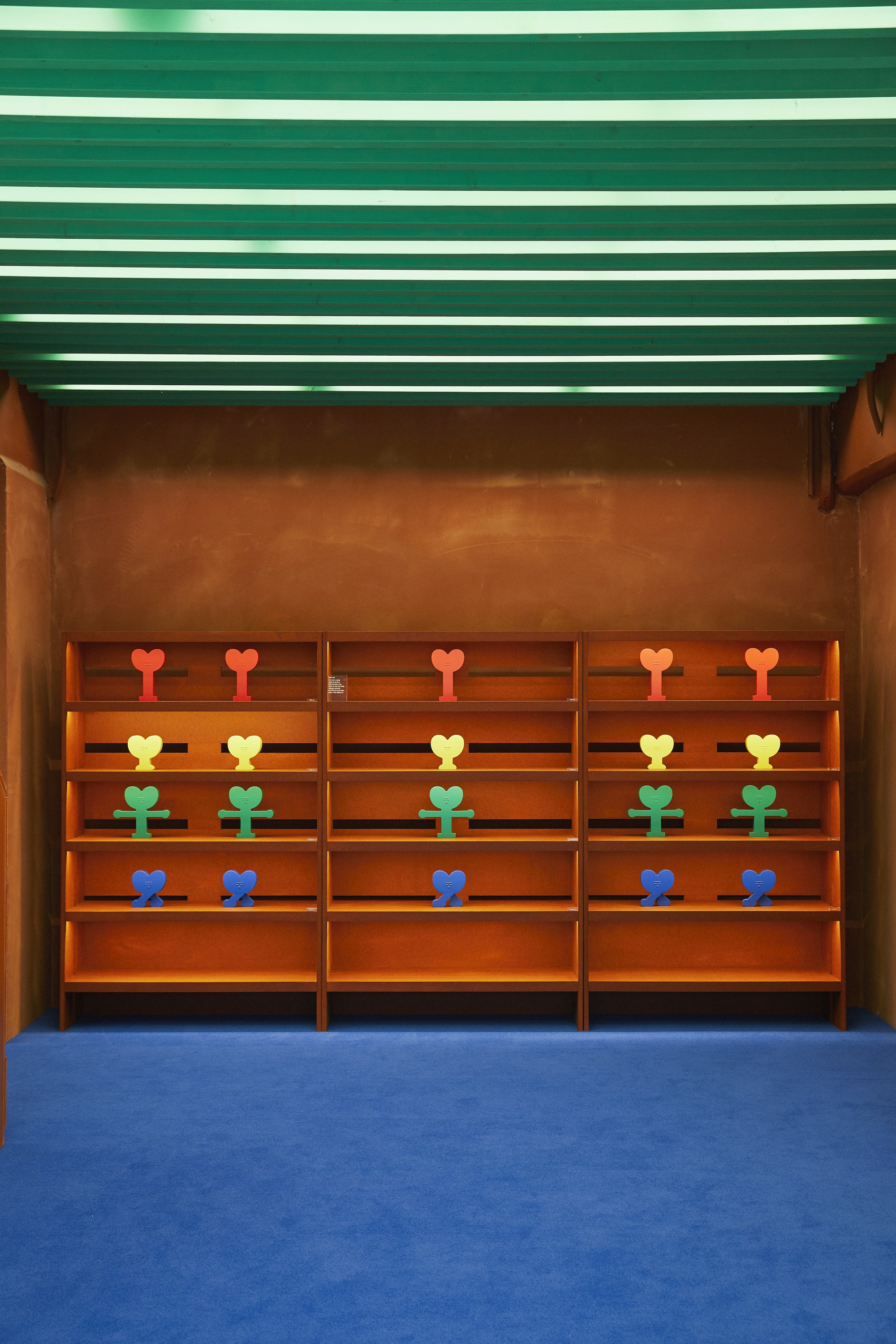 Built-in shelving wall displaying colorful sculptural objects in rows beneath green linear ceiling strips