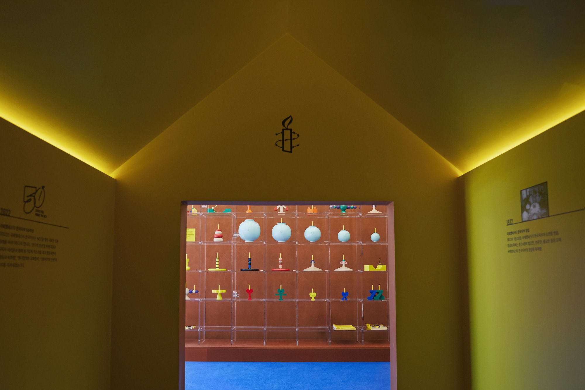 Doorway framing a display vitrine with suspended objects, yellow cove lighting illuminating the gabled ceiling