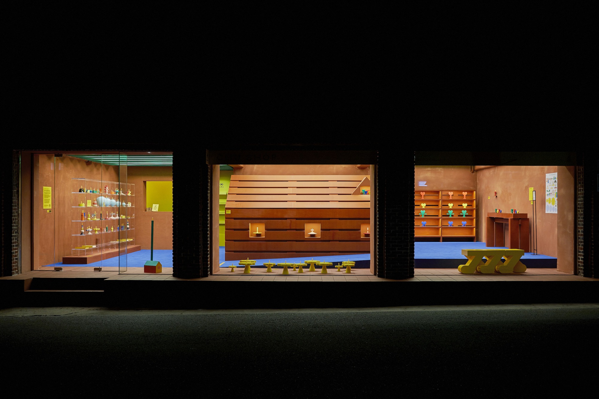 Three storefronts at night revealing interiors with timber furnishings, shelving, and blue flooring