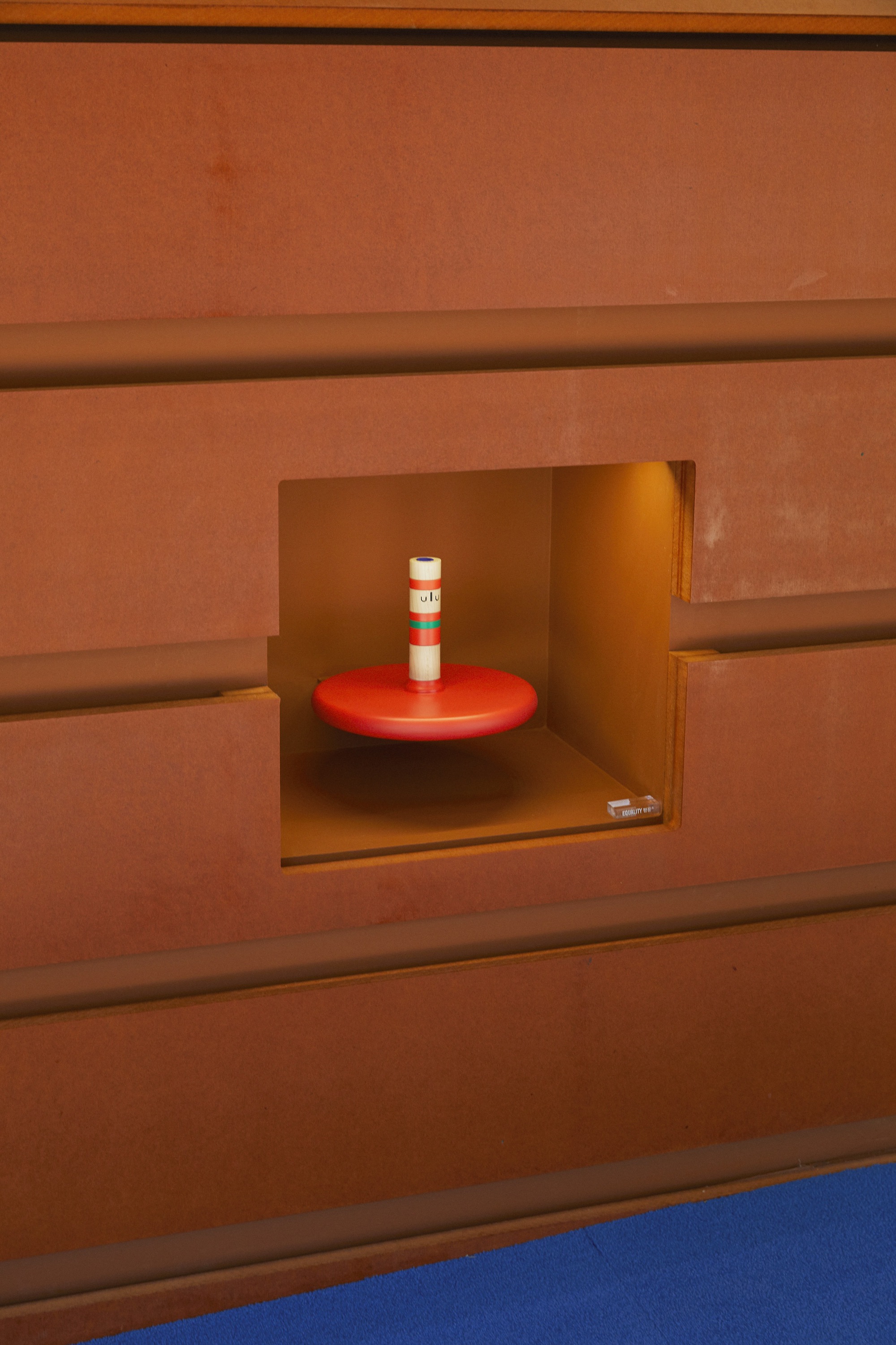 Square niche in terracotta-toned wall with a red round shelf and white candle inside