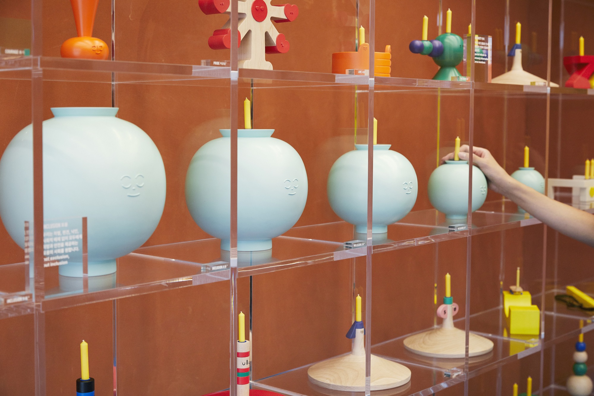 Transparent acrylic shelving displaying white ceramic vessels and candleholders with a hand reaching in