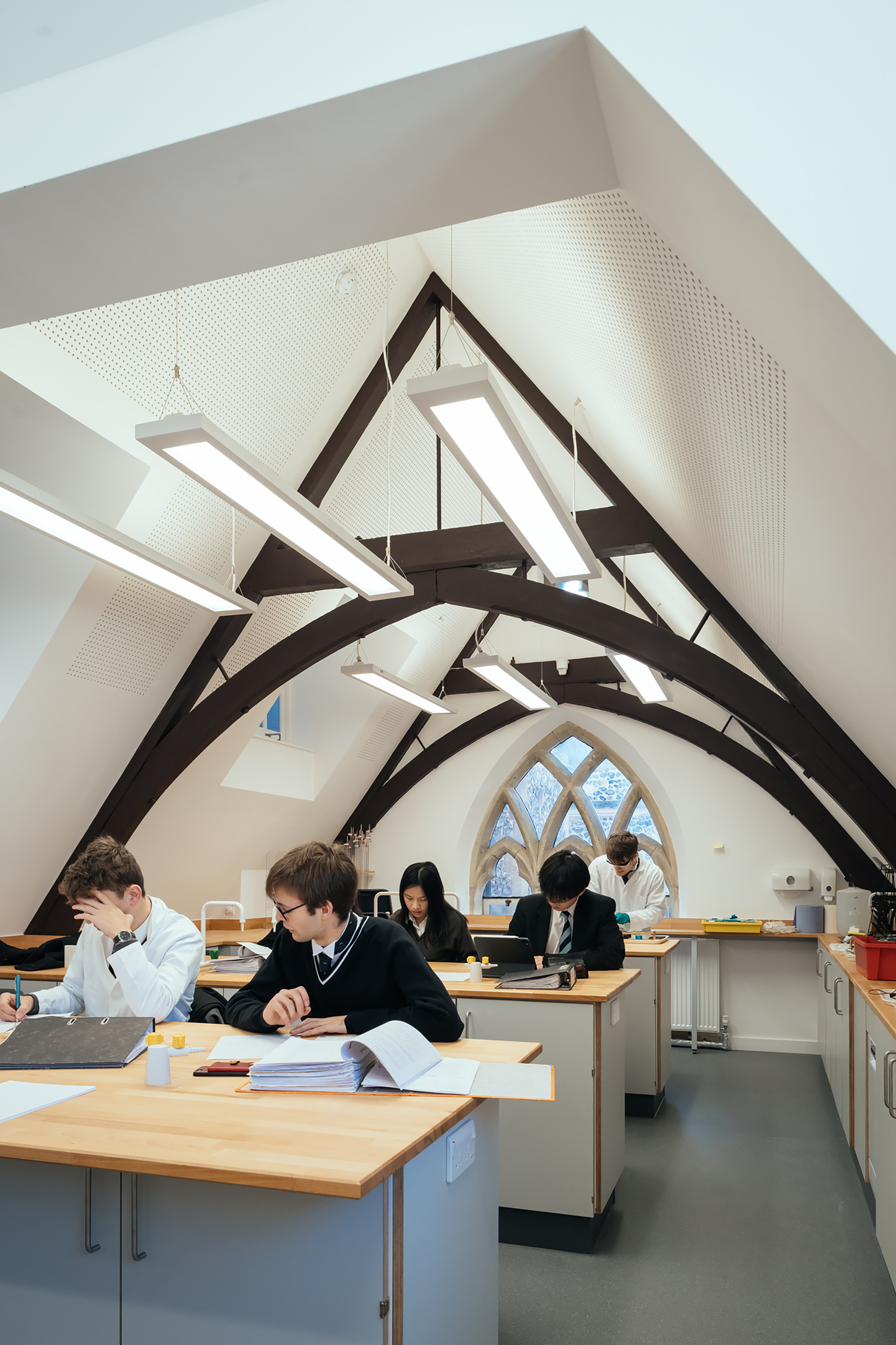 Interior space with exposed timber trusses and a gothic arched window where students work at benches